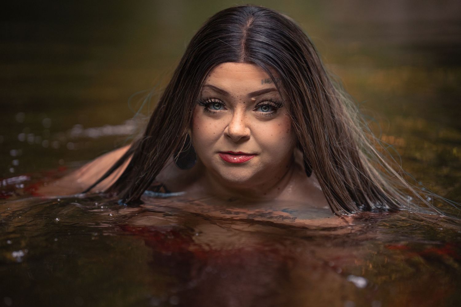 Woman with long wet hair submerged in natural water, gazing at camera with dramatic makeup.