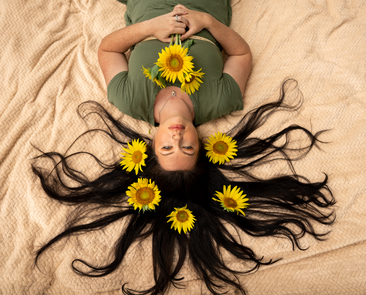 Woman lying on bed with long black hair fanned out, adorned with yellow sunflowers, wearing a green top.
