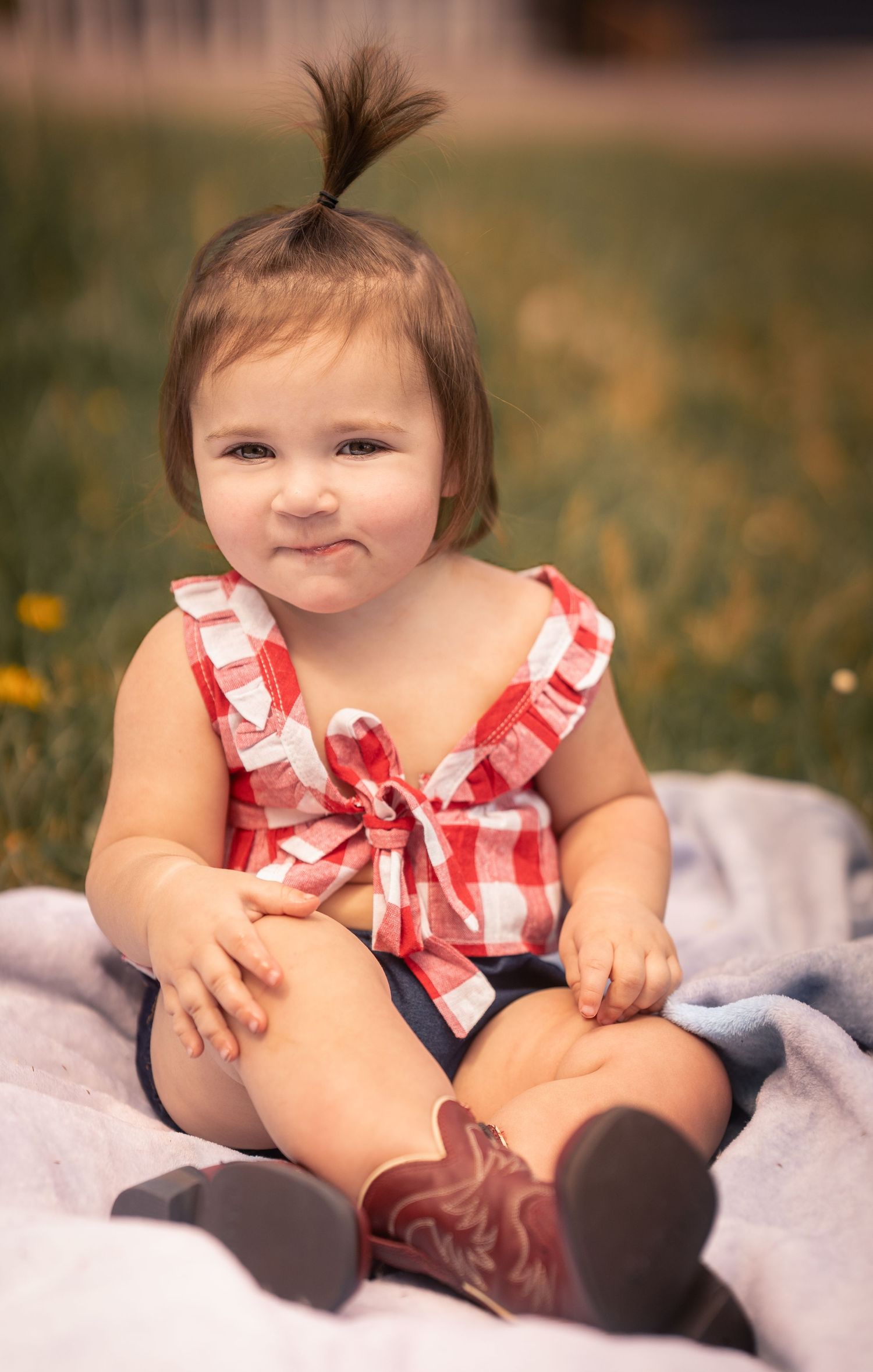Adorable baby girl in red plaid top and cowboy boots sitting on blanket outdoors smiling.