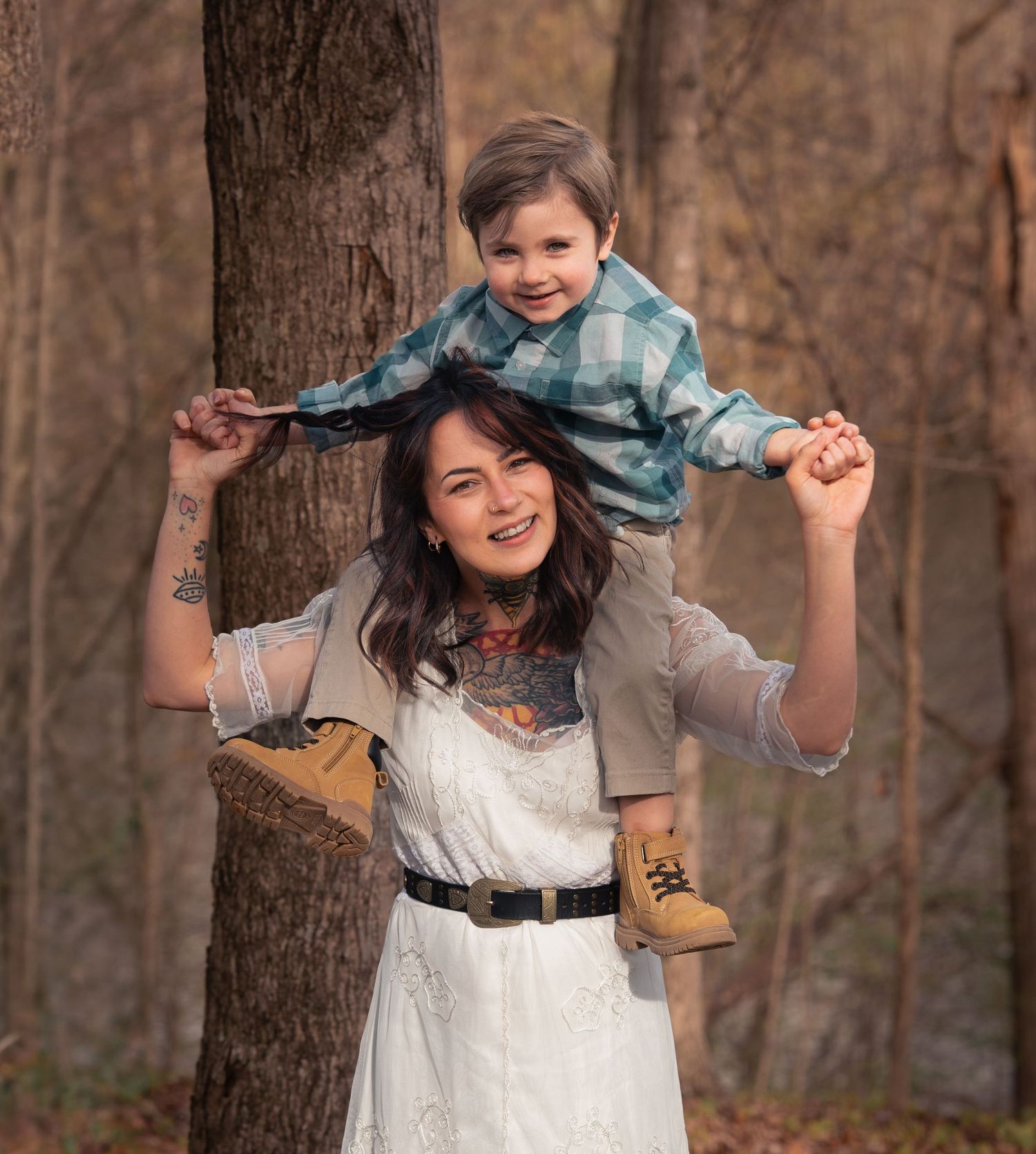 Woman in white dress smiles with young boy sitting on her shoulders in an autumn forest setting.