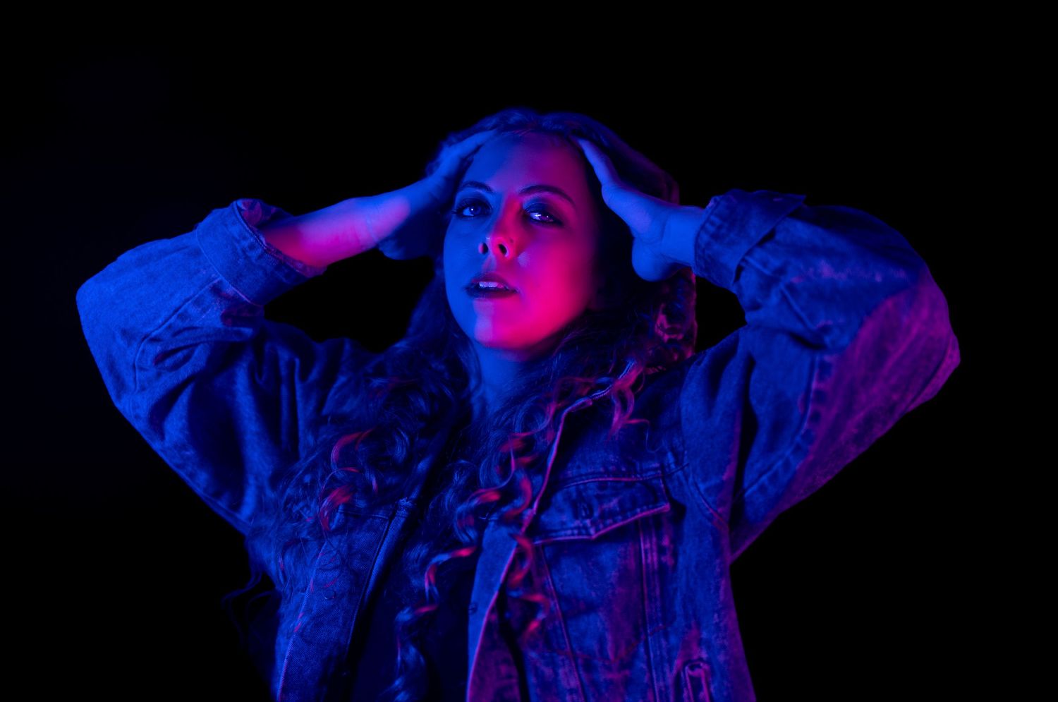Woman in denim jacket poses dramatically under neon blue and red lights against dark background.