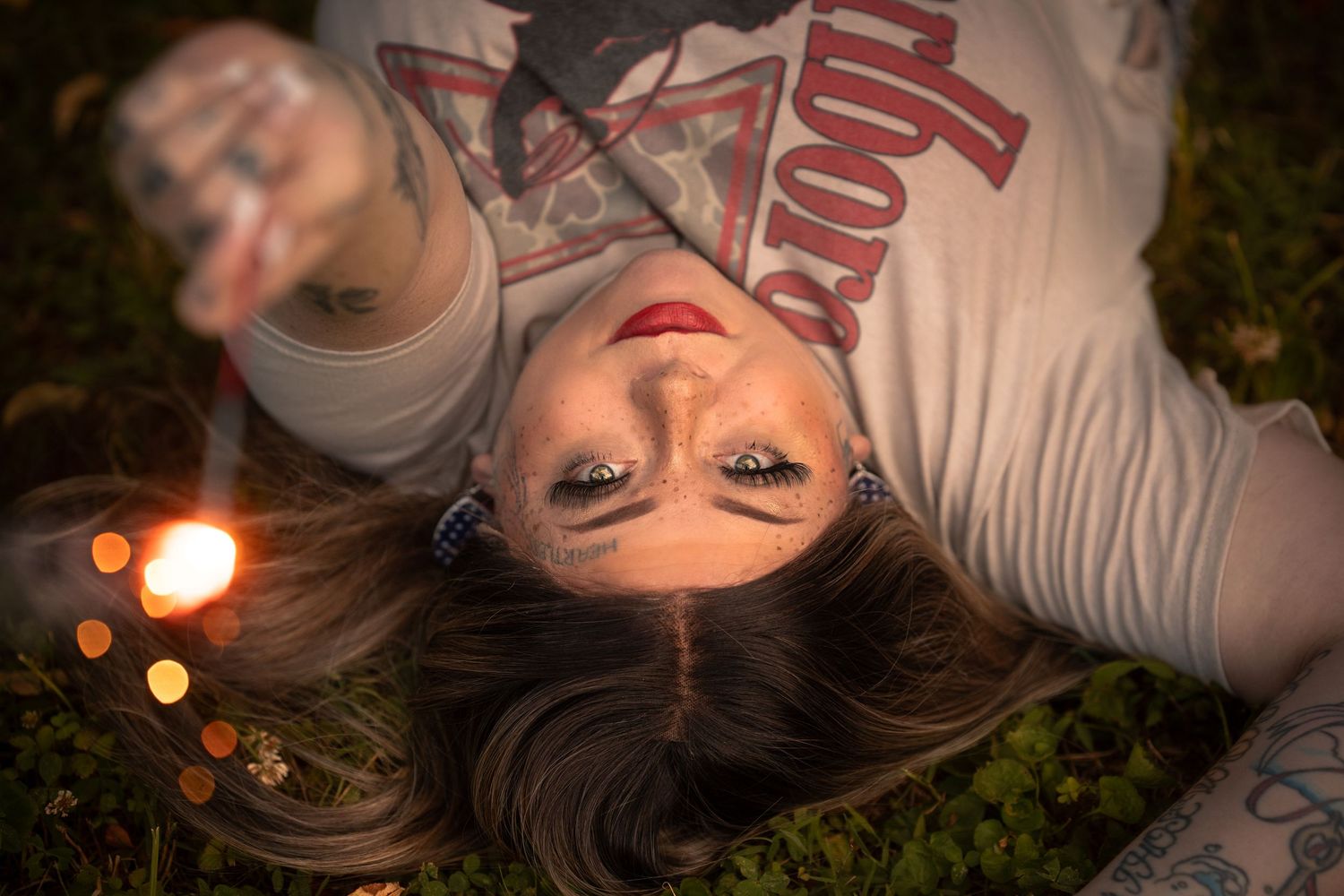Woman with red lips and long lashes lying on grass, holding sparkler, wearing Marlboro tee with tattoos visible.