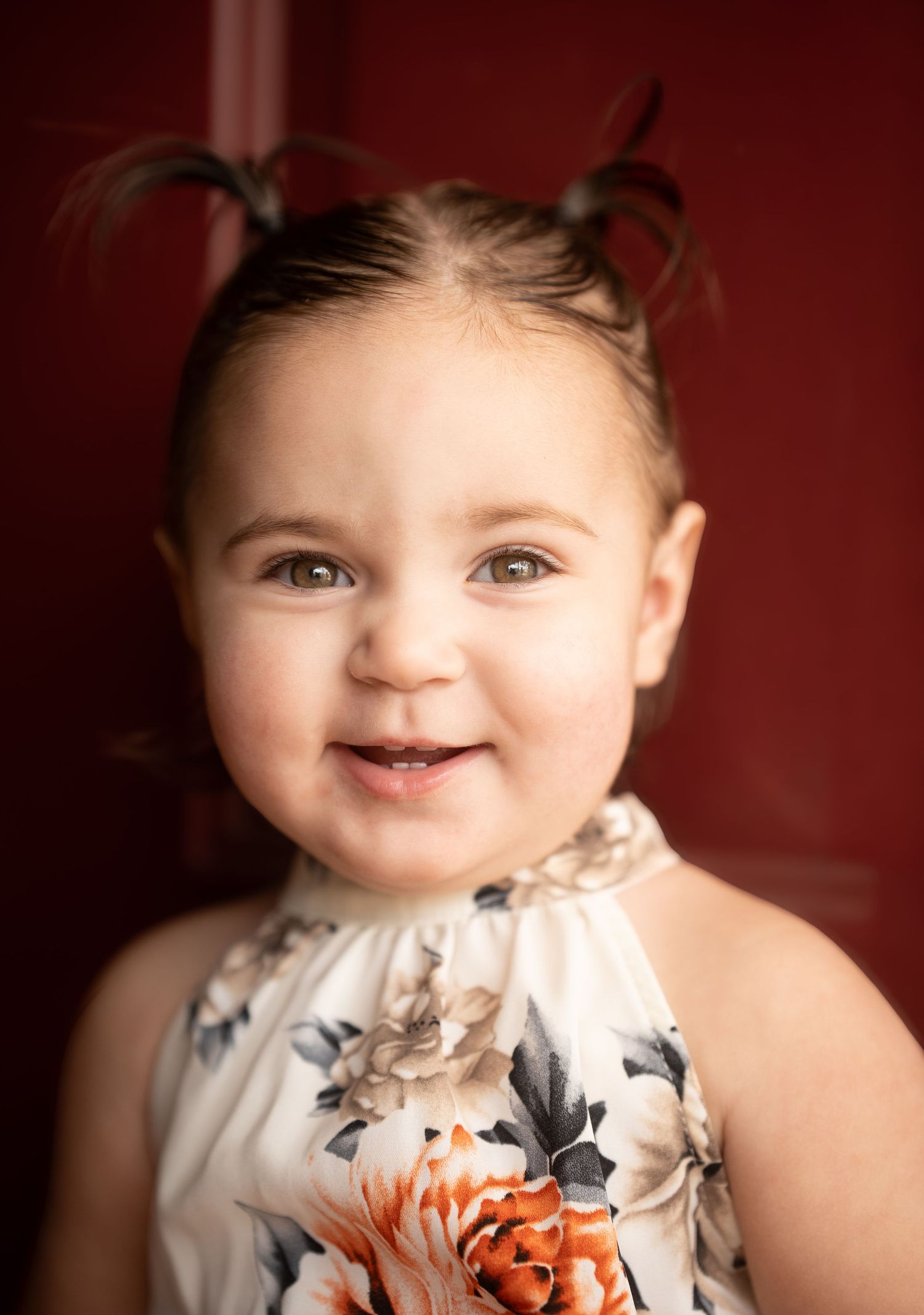 Smiling toddler girl with pigtails wearing a floral dress poses against a red background.