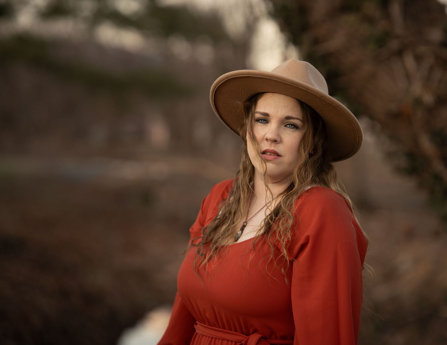 Woman in red dress and tan hat poses outdoors with blurred natural background.