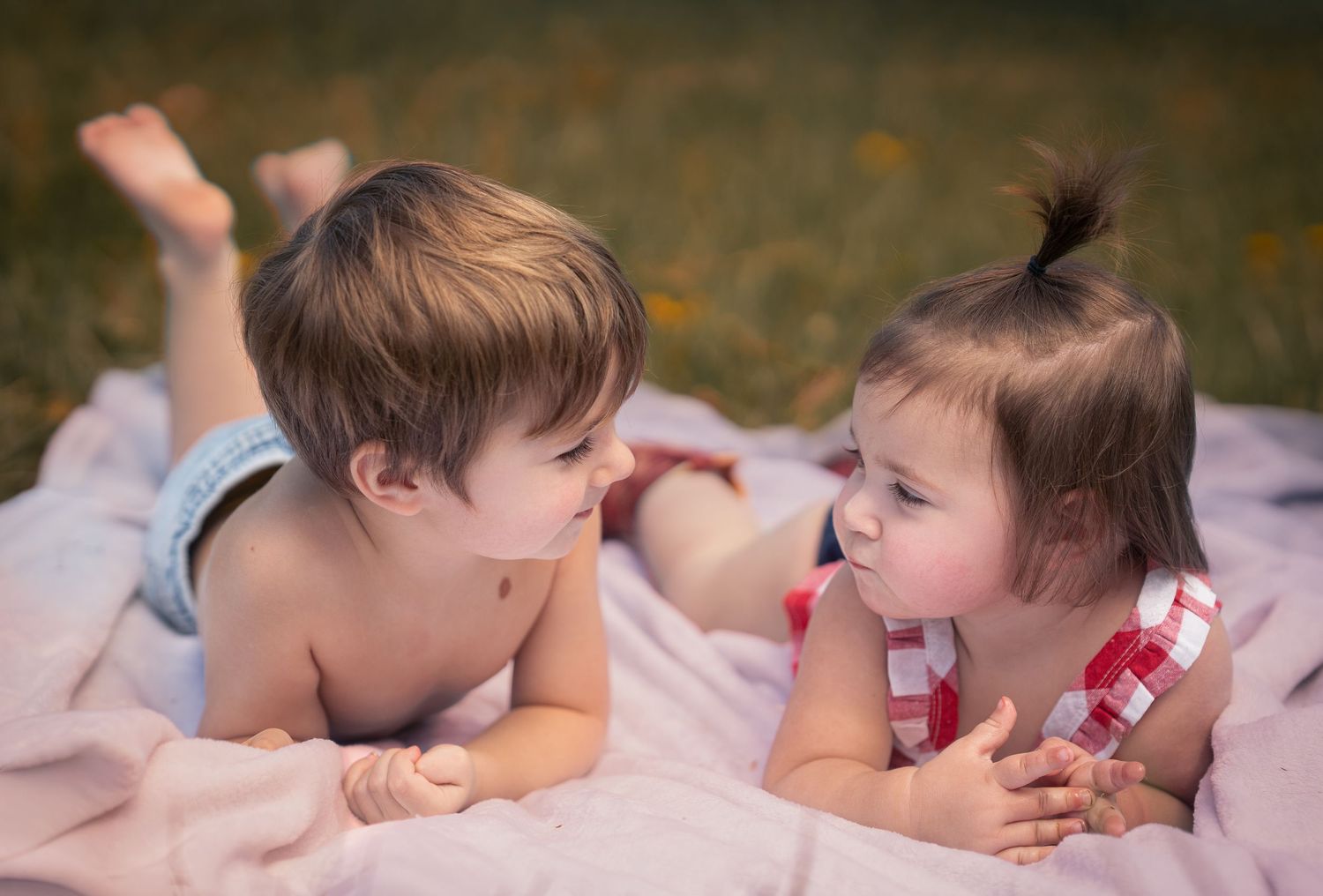 Two adorable toddlers lying on a pink blanket outdoors, facing each other in a sweet moment of childhood friendship.