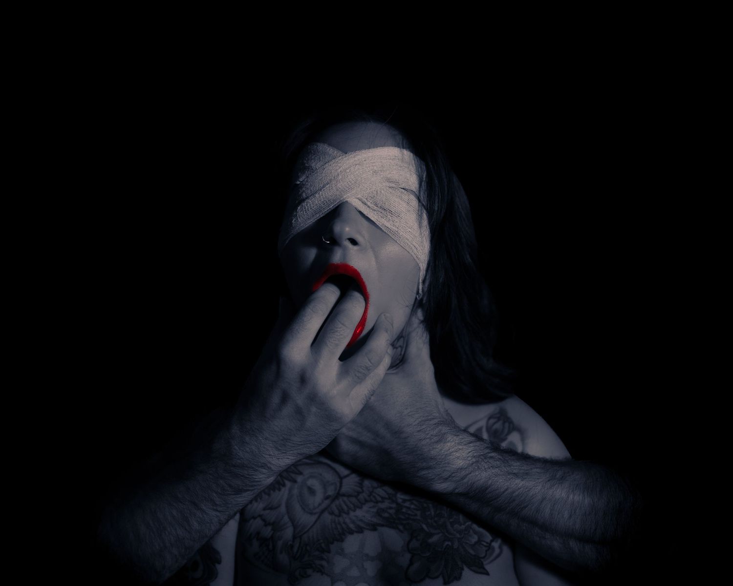 Dark artistic photo of a person with bandaged face, red lips, and tattooed arms in dramatic horror-themed portrait.