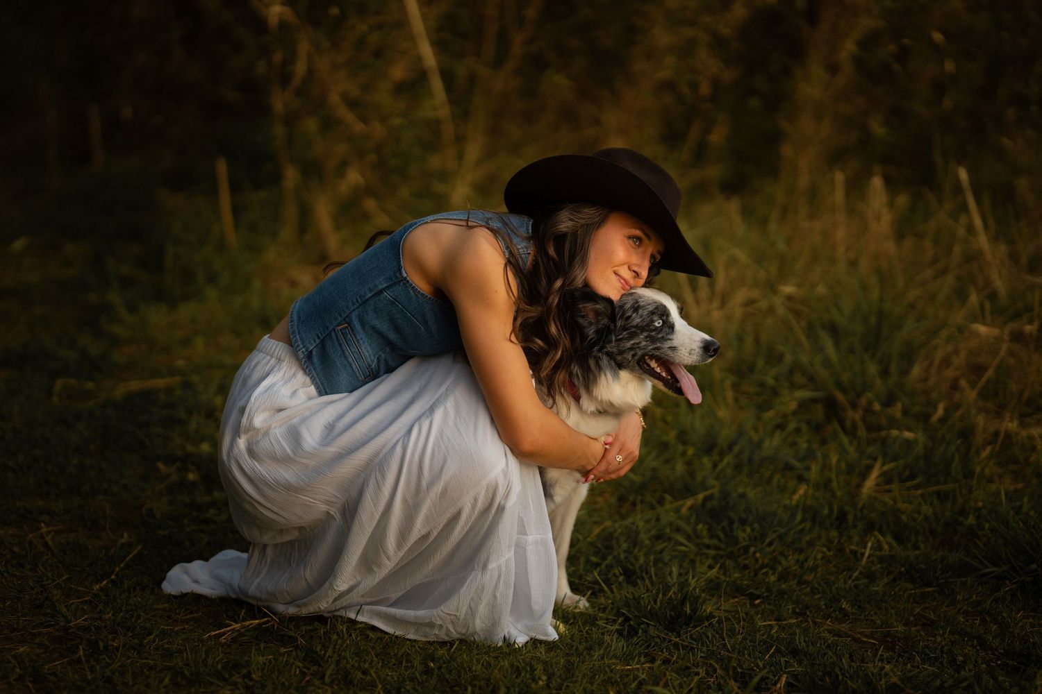 Woman in cowboy hat and white skirt tenderly embracing Australian Shepherd dog outdoors.