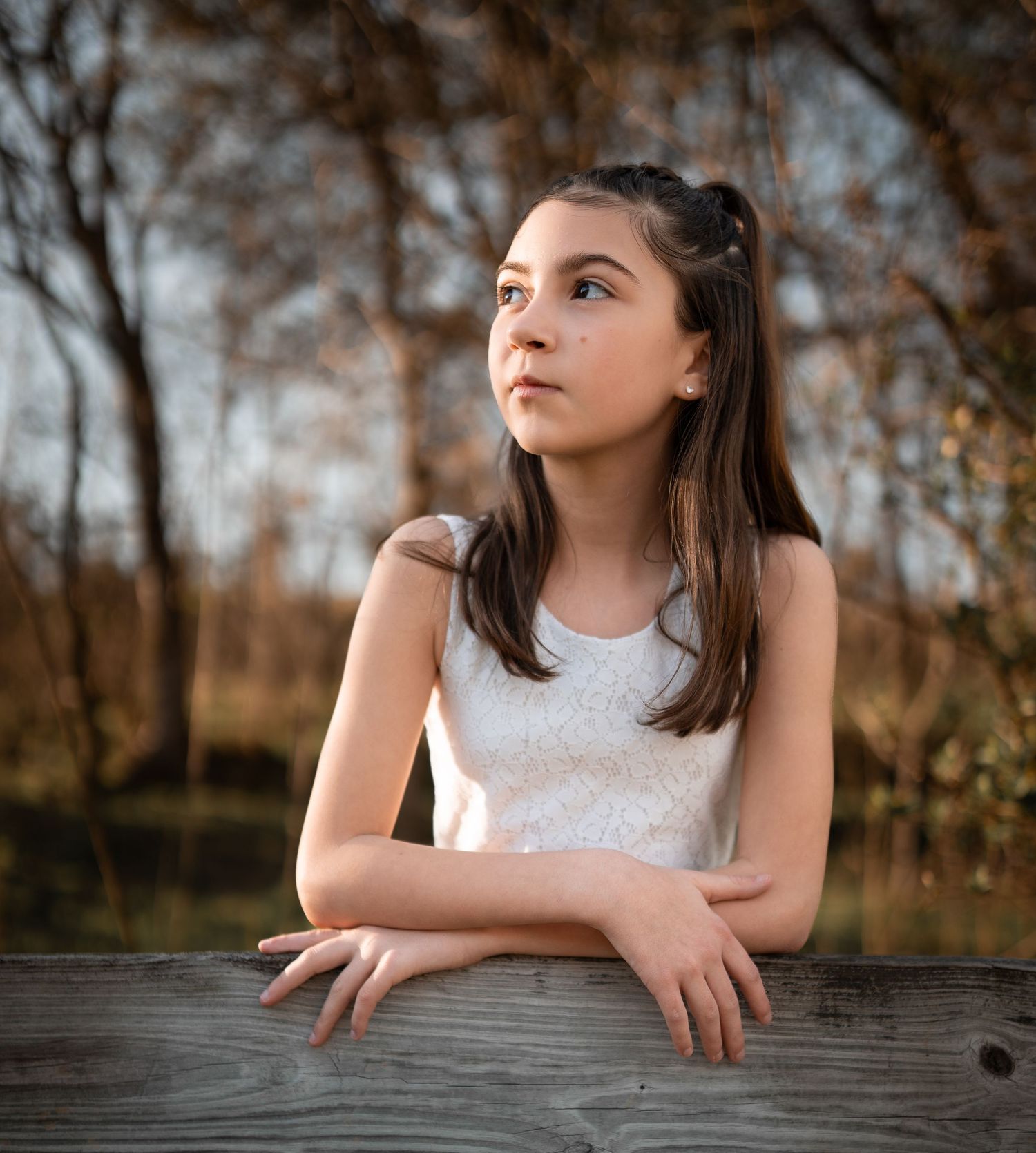 Young girl in white lace top leans on wooden fence gazing thoughtfully outdoors.