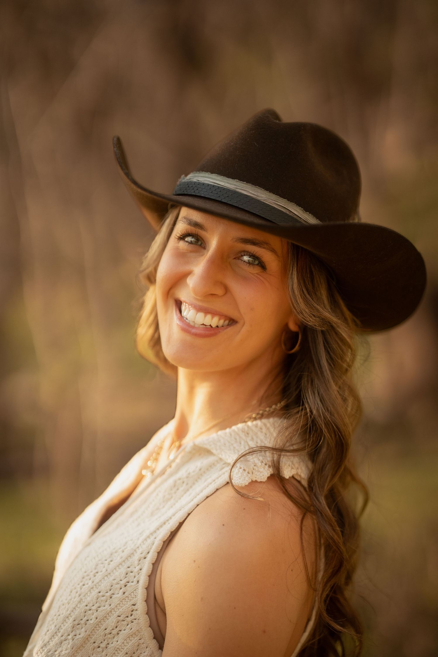 Smiling woman with wavy hair wearing black cowboy hat in warm golden sunset light.