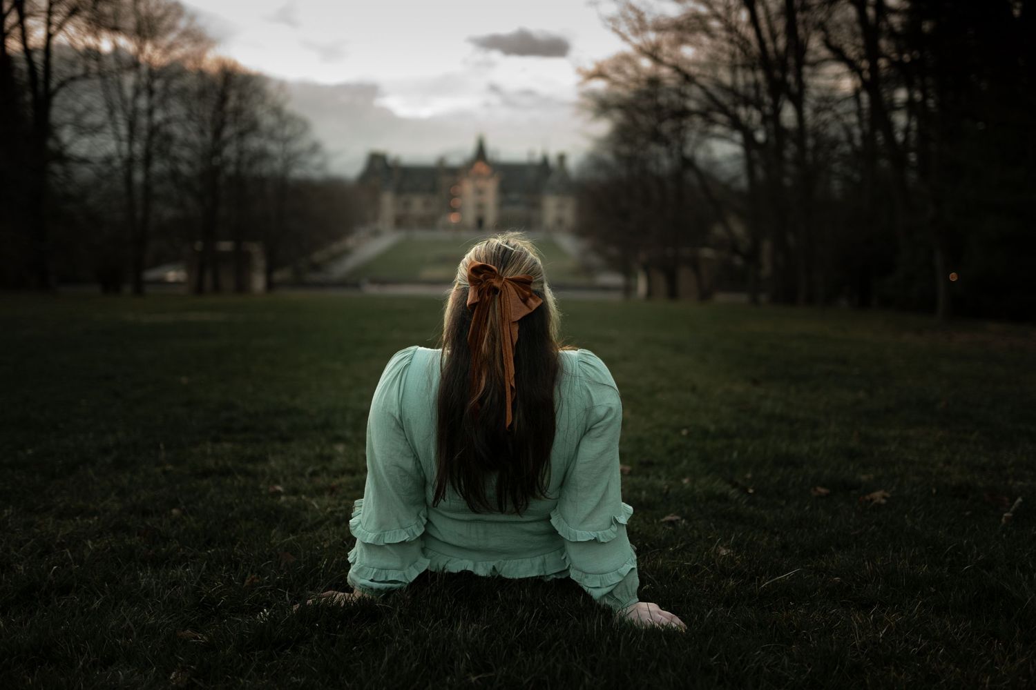 Young woman in mint blouse sits on lush green grass facing a grand historic mansion at dusk.