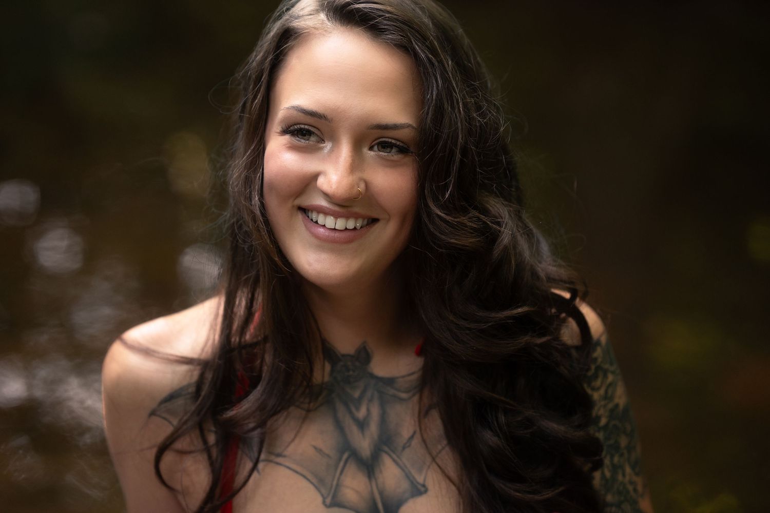Smiling young woman with long dark hair and chest tattoo posing outdoors near water.