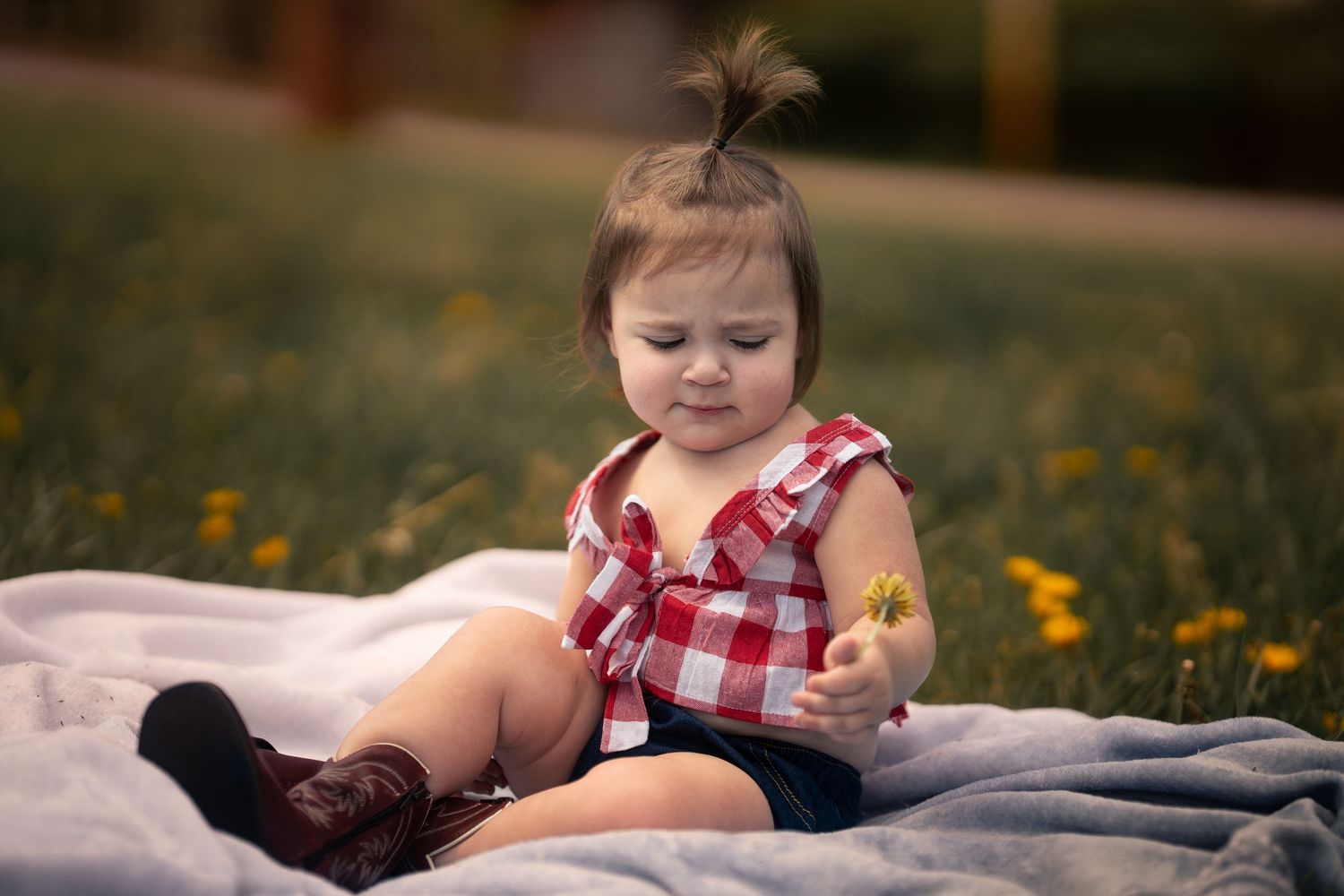 Adorable baby girl in red plaid top sitting on blanket in green field, holding a yellow dandelion flower.
