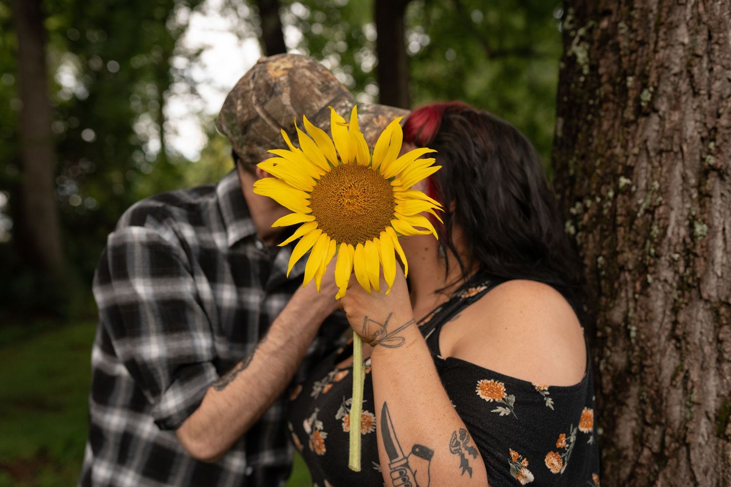 Couple hiding behind a large sunflower near a tree in a romantic outdoor photo shoot.