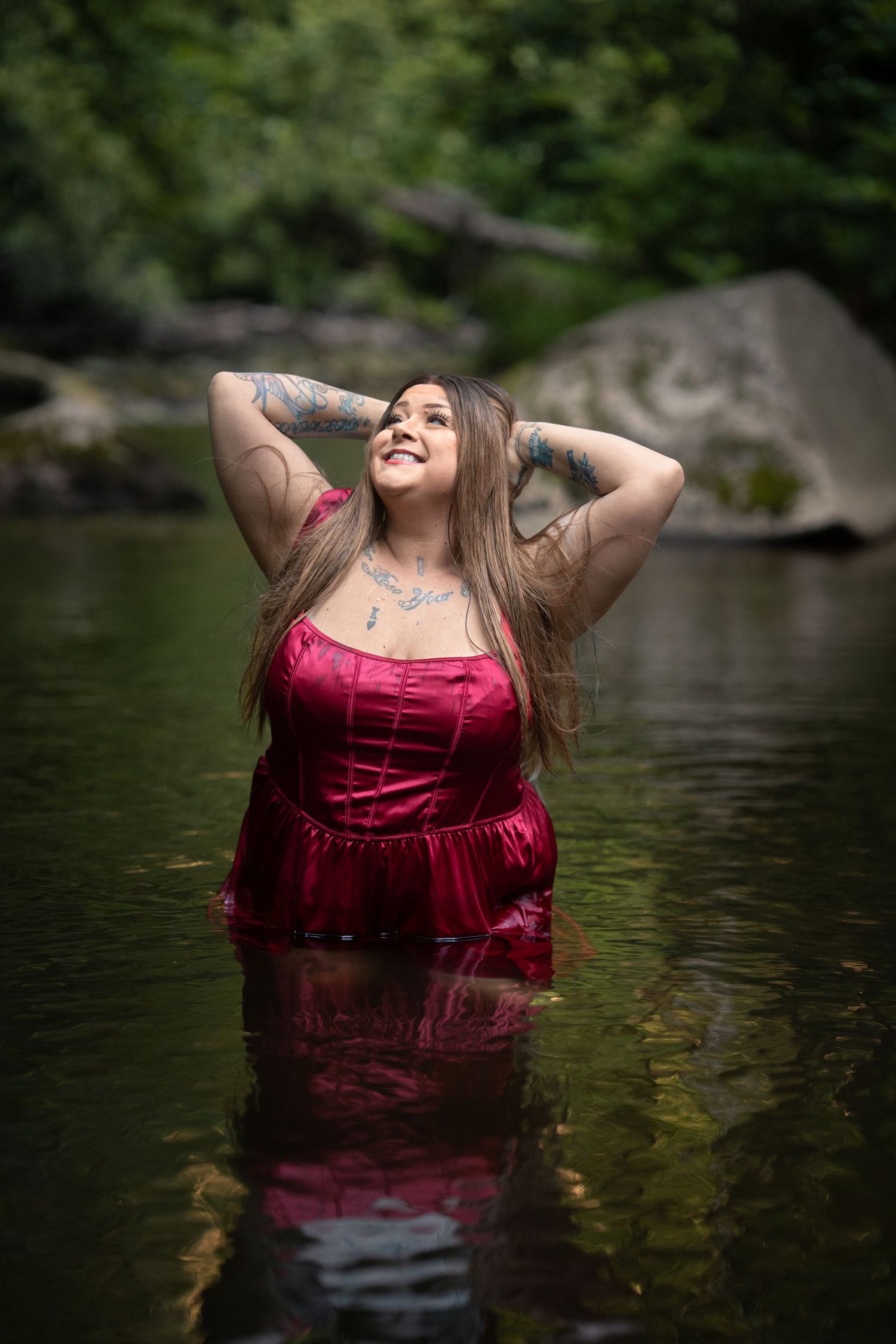 A tattooed woman in a red satin dress stands waist-deep in a serene forest river, smiling with hands behind her head.