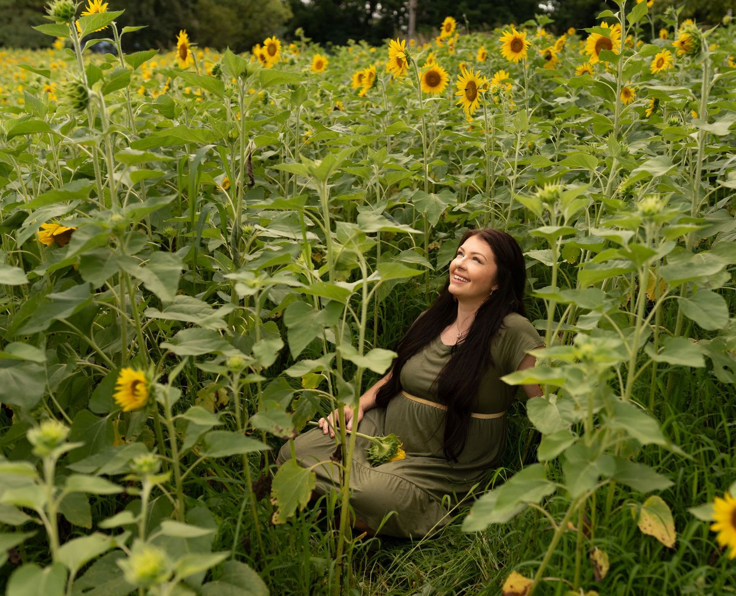 Woman in olive green outfit sitting among tall sunflowers in a lush green field, smiling joyfully.