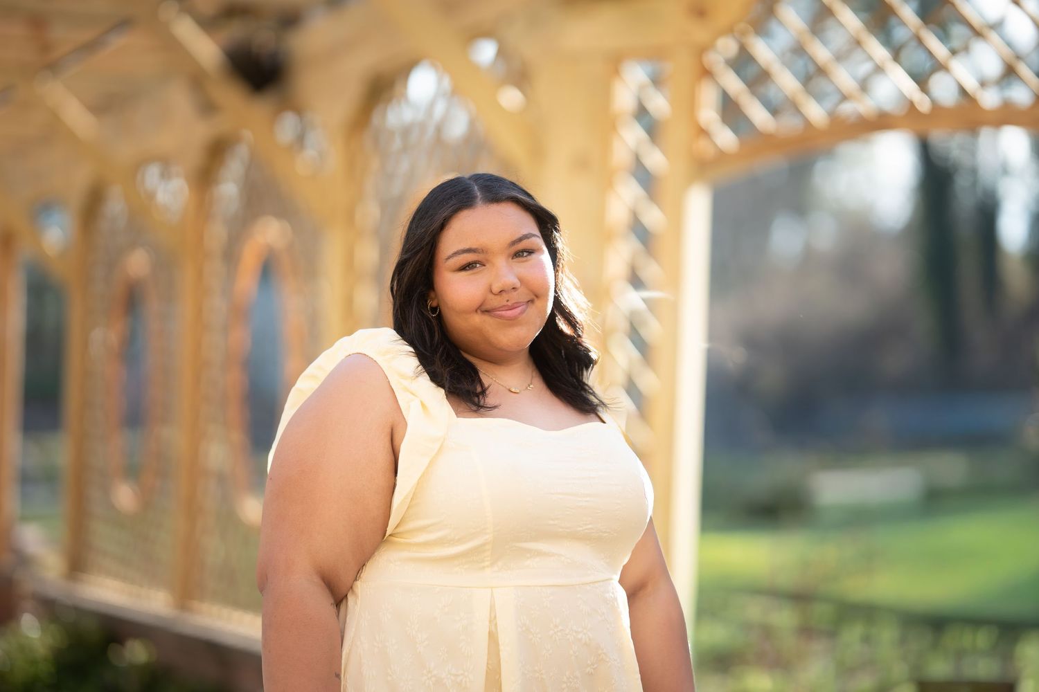 Young woman in yellow sundress smiling under decorative garden pergola in bright daylight.