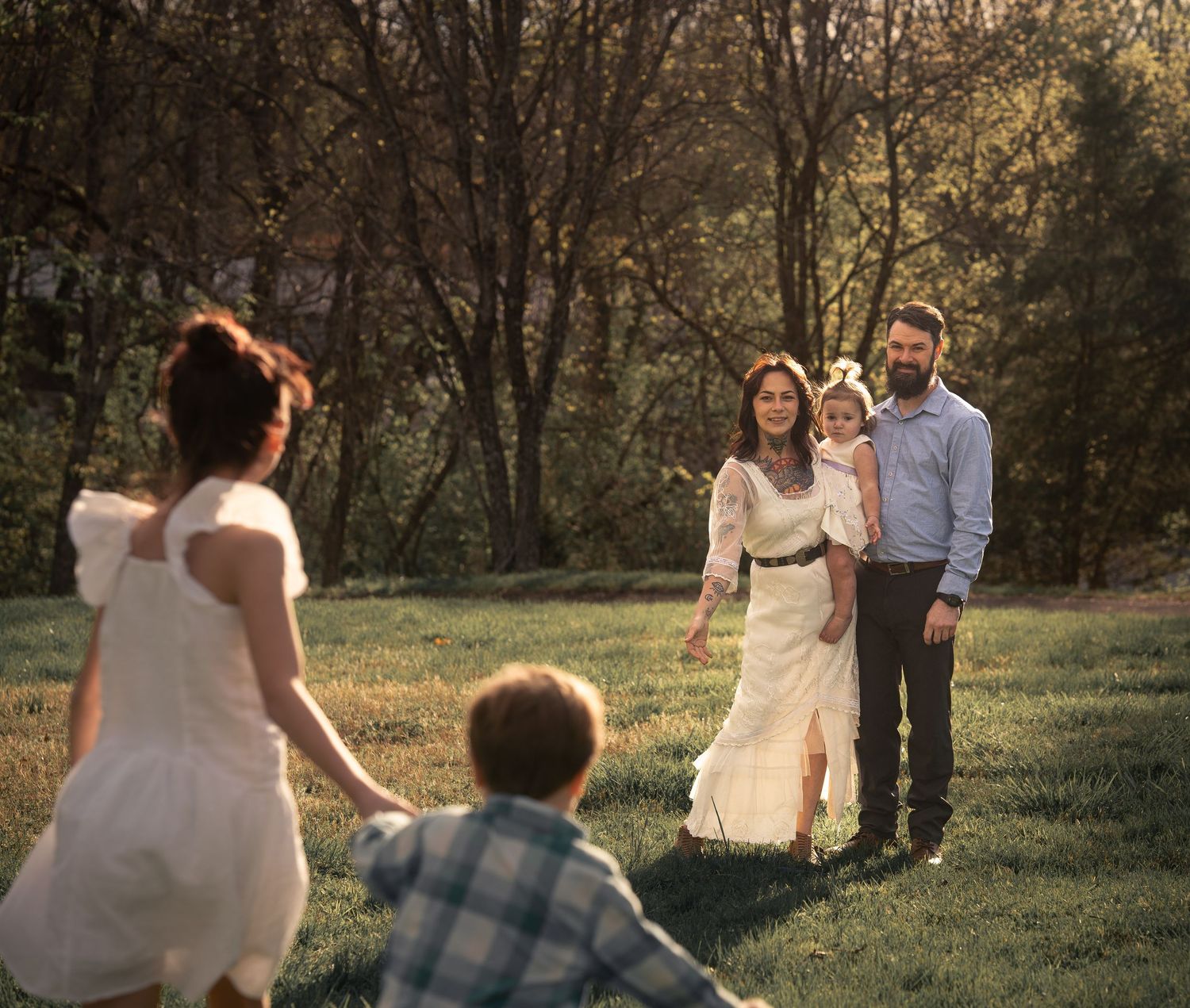 Family portrait session outdoors with parents holding child while two kids run toward them on green lawn.