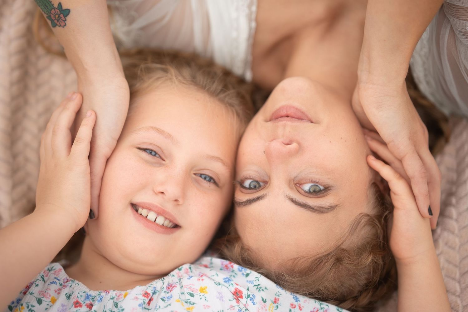 A woman and young girl lying down together, smiling warmly, with hands gently framing each other's faces.