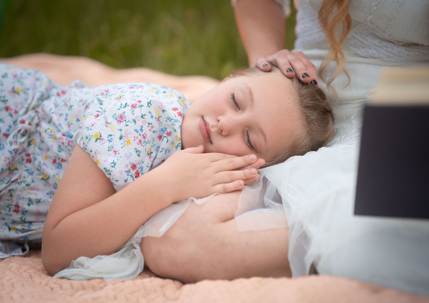 Young girl sleeping peacefully outdoors on a pink blanket while a woman gently strokes her hair.