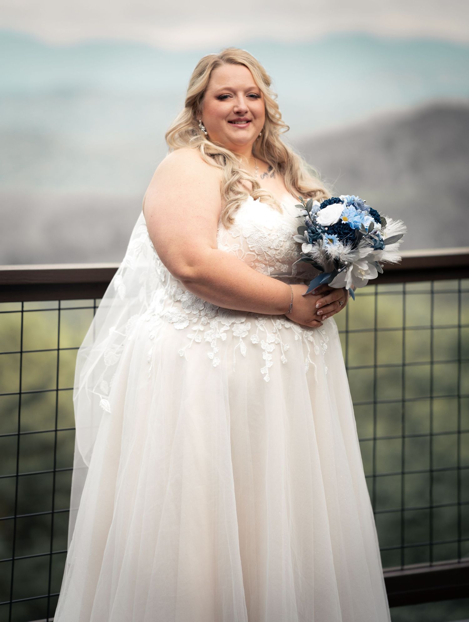 Bride in white lace tulle gown holding blue floral bouquet, posing on outdoor deck with mountain views.