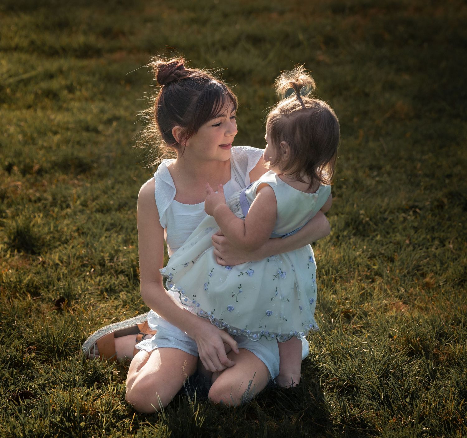 A young girl kneeling on green grass, tenderly embracing her toddler sister in matching light summer dresses.