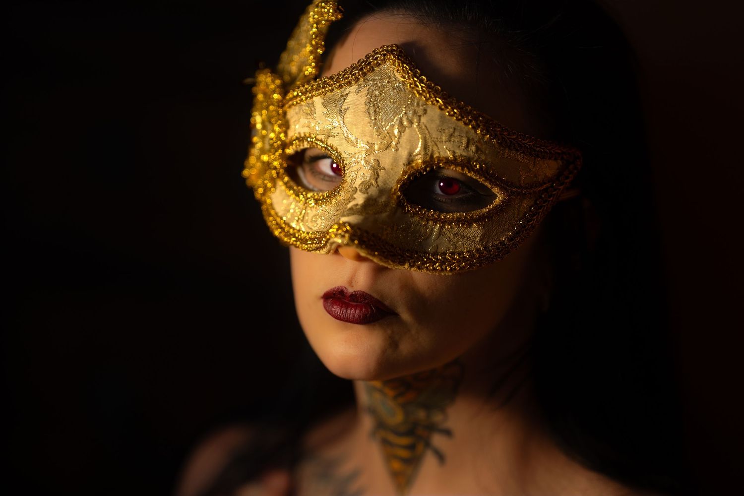 Close-up portrait of a woman wearing a gold masquerade mask with neck tattoos and red lips.