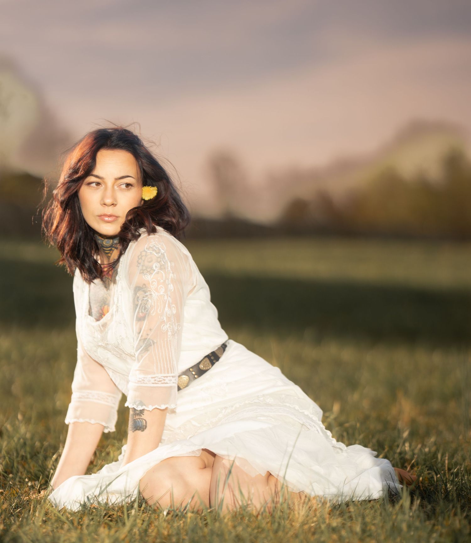 Woman in white boho dress sitting in a sunlit green meadow with a yellow flower.