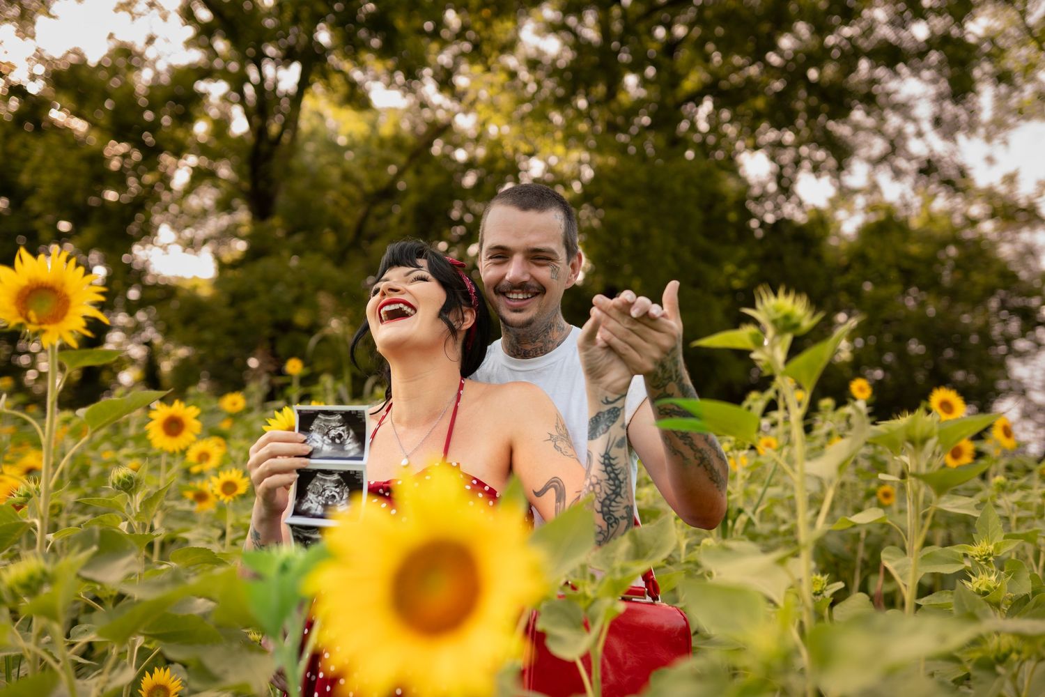 Happy couple in sunflower field holding ultrasound photo announcing pregnancy, surrounded by blooming sunflowers and trees.