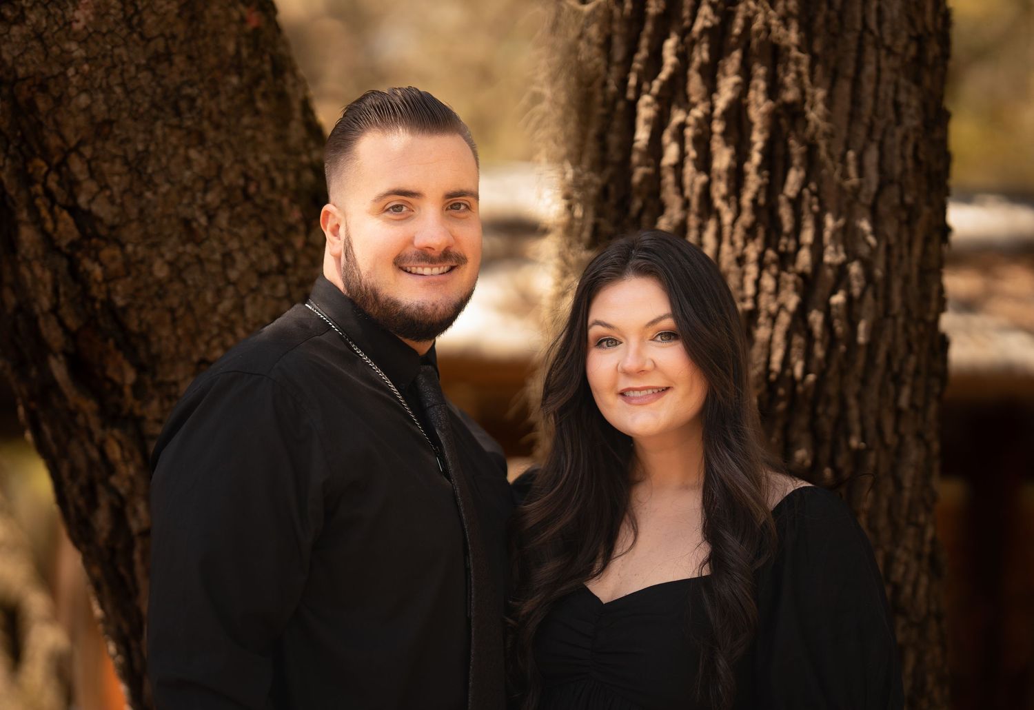 A couple in matching black outfits smiling together in front of a large tree in autumn.
