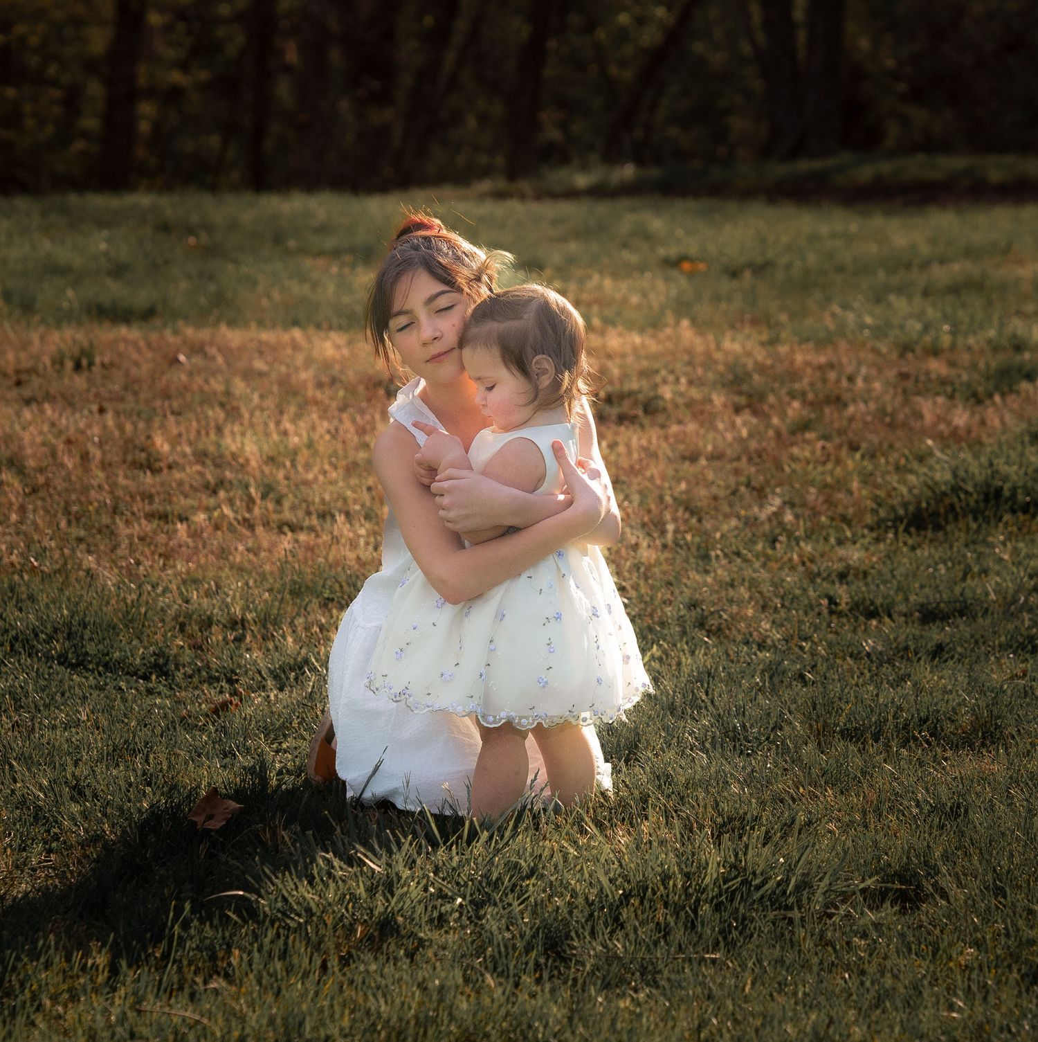 Older girl in white dress hugging toddler sibling on green grass in warm golden sunset light.