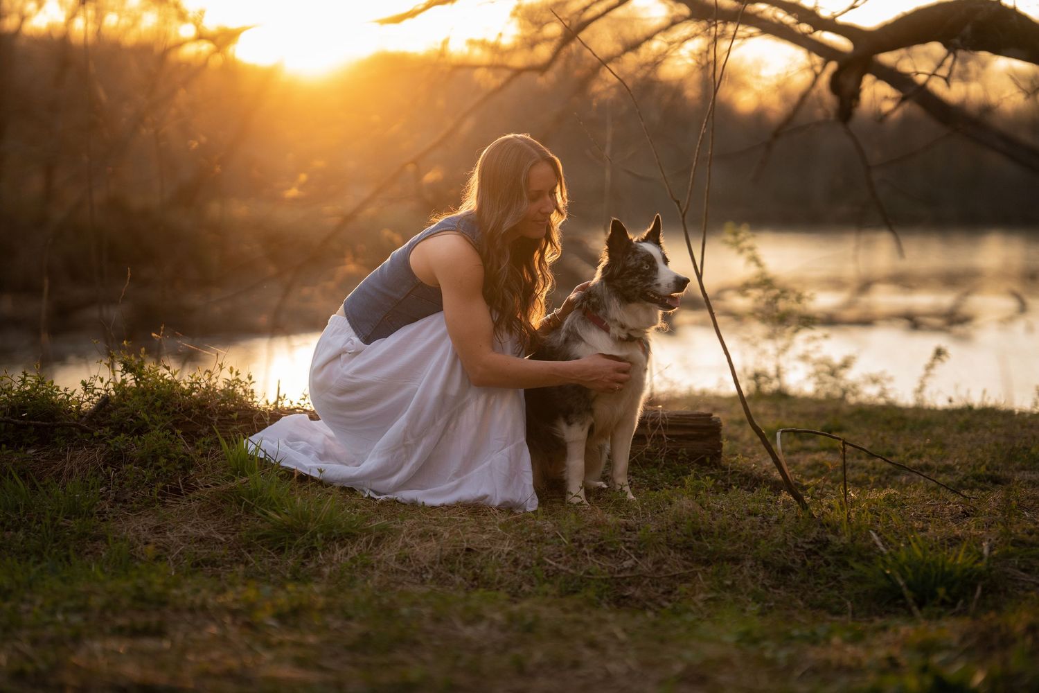 Young woman in white dress cuddles with husky dog by lake at sunset.
