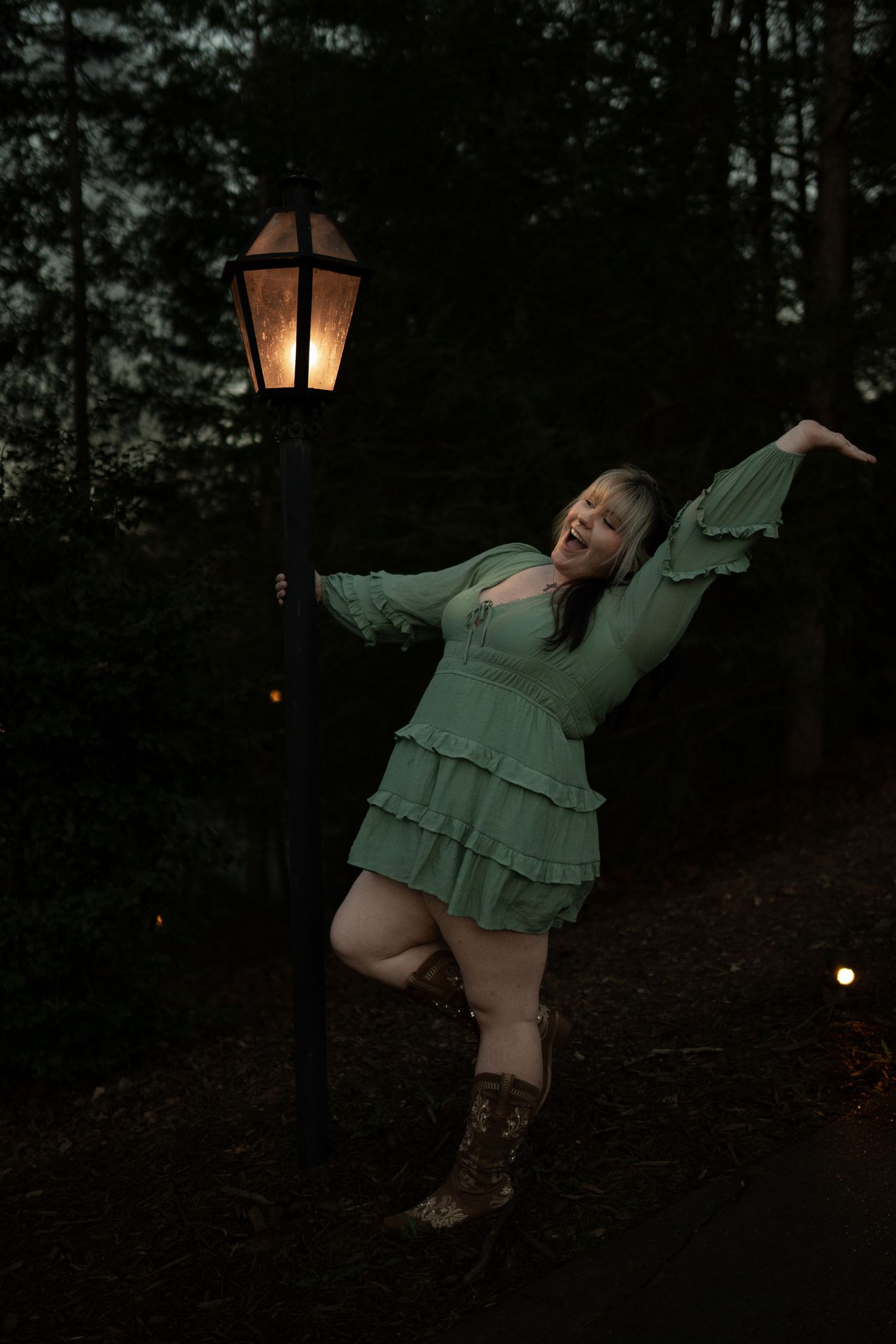 Woman in green tiered mini dress dancing joyfully beside glowing lamppost at dusk in forest.