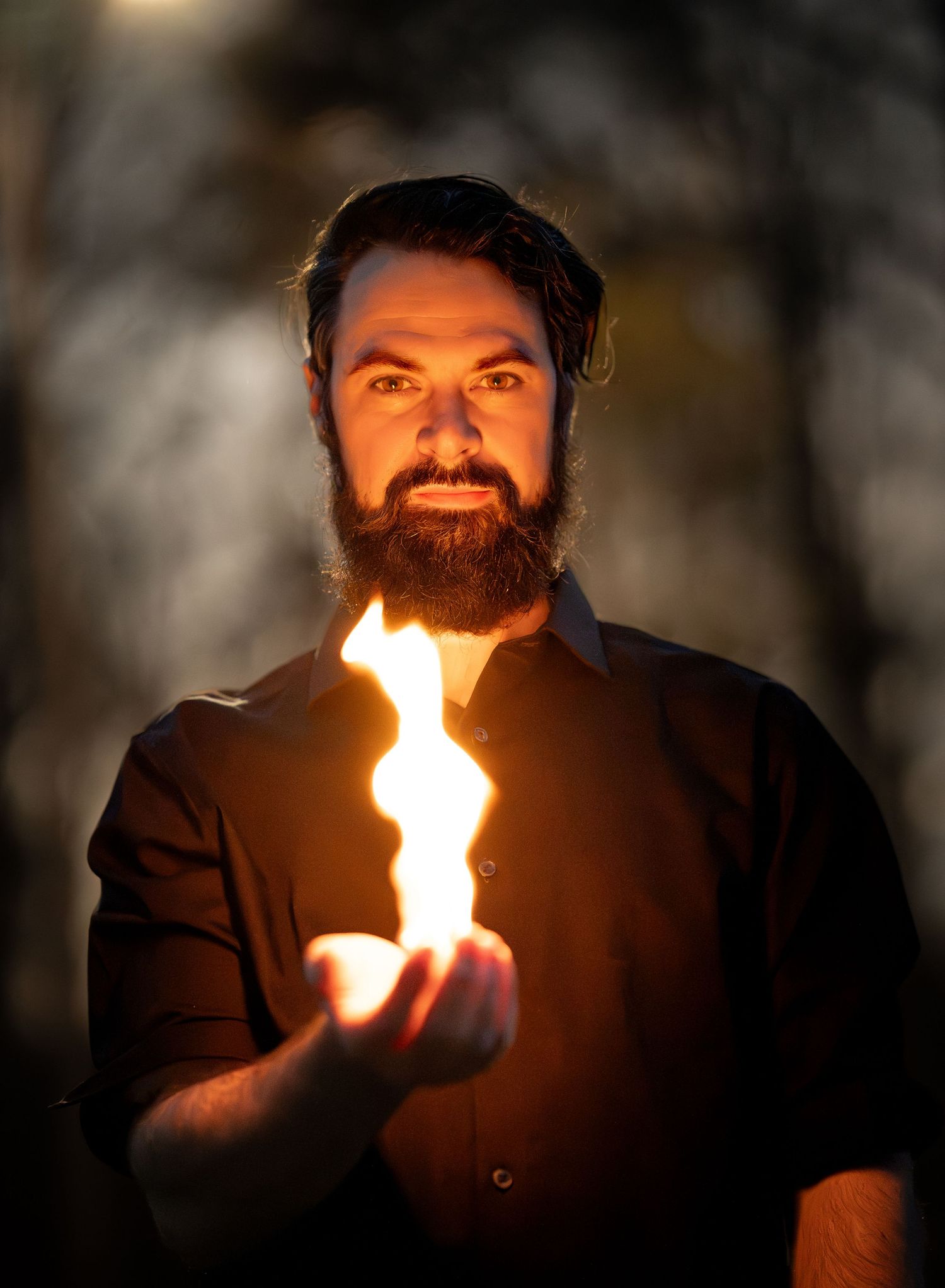 Bearded man in black shirt holds flame in palm during dramatic outdoor portrait at dusk.