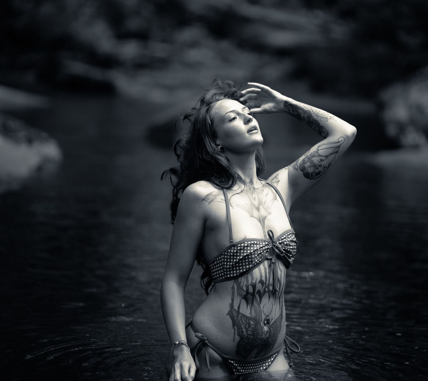 Black and white photo of a tattooed woman standing in water, head tilted back, hand in hair.