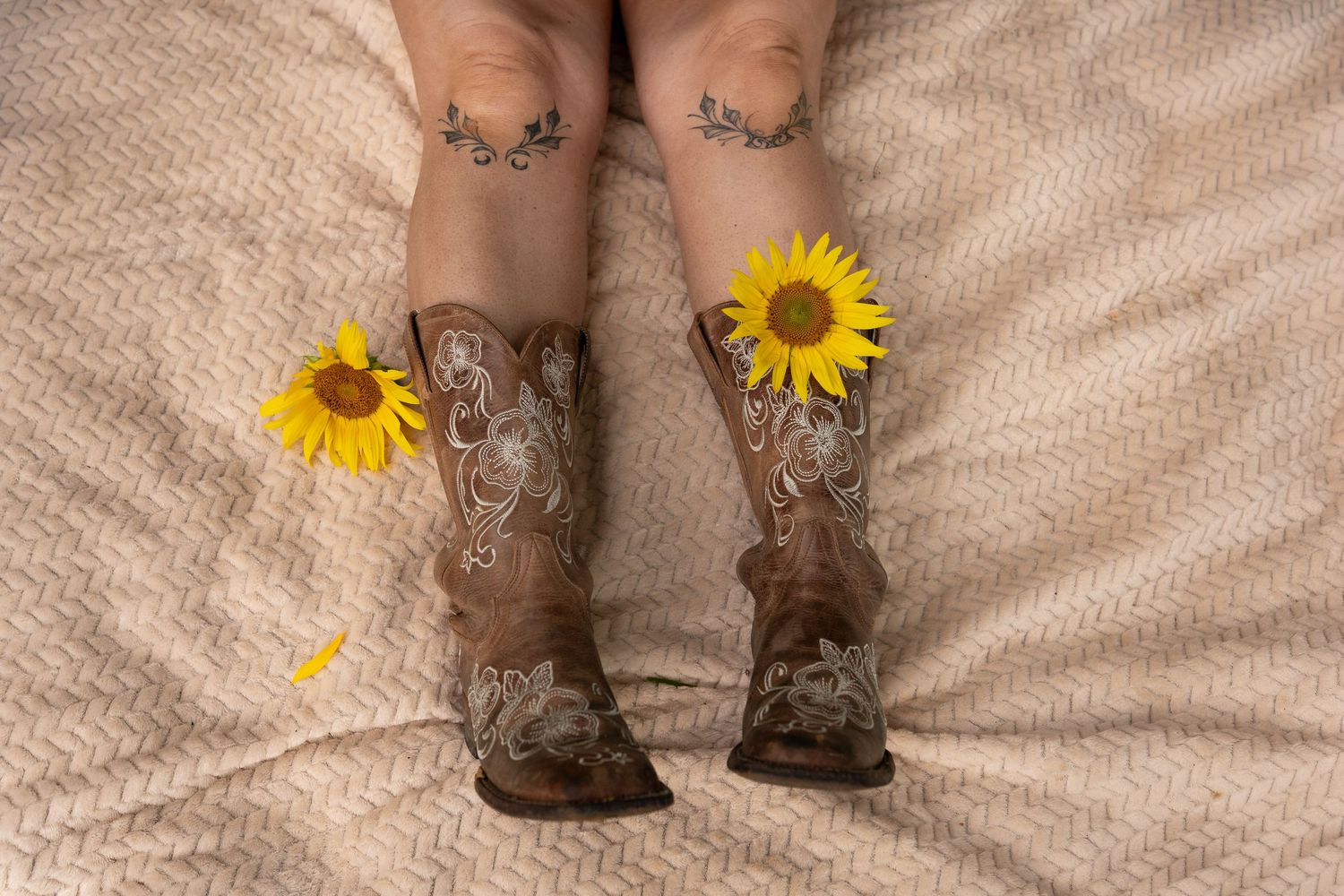 Person wearing brown cowboy boots with sunflowers tucked inside, sitting on a beige textured blanket with floral leg tattoos visible.