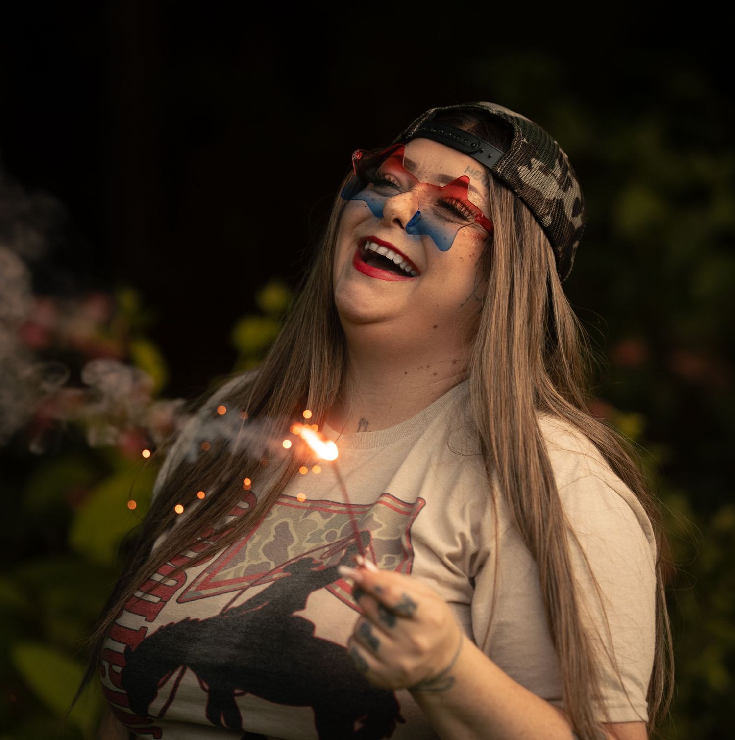 Woman laughing while holding a sparkler outdoors, wearing camo hat and patriotic face paint at a celebration.