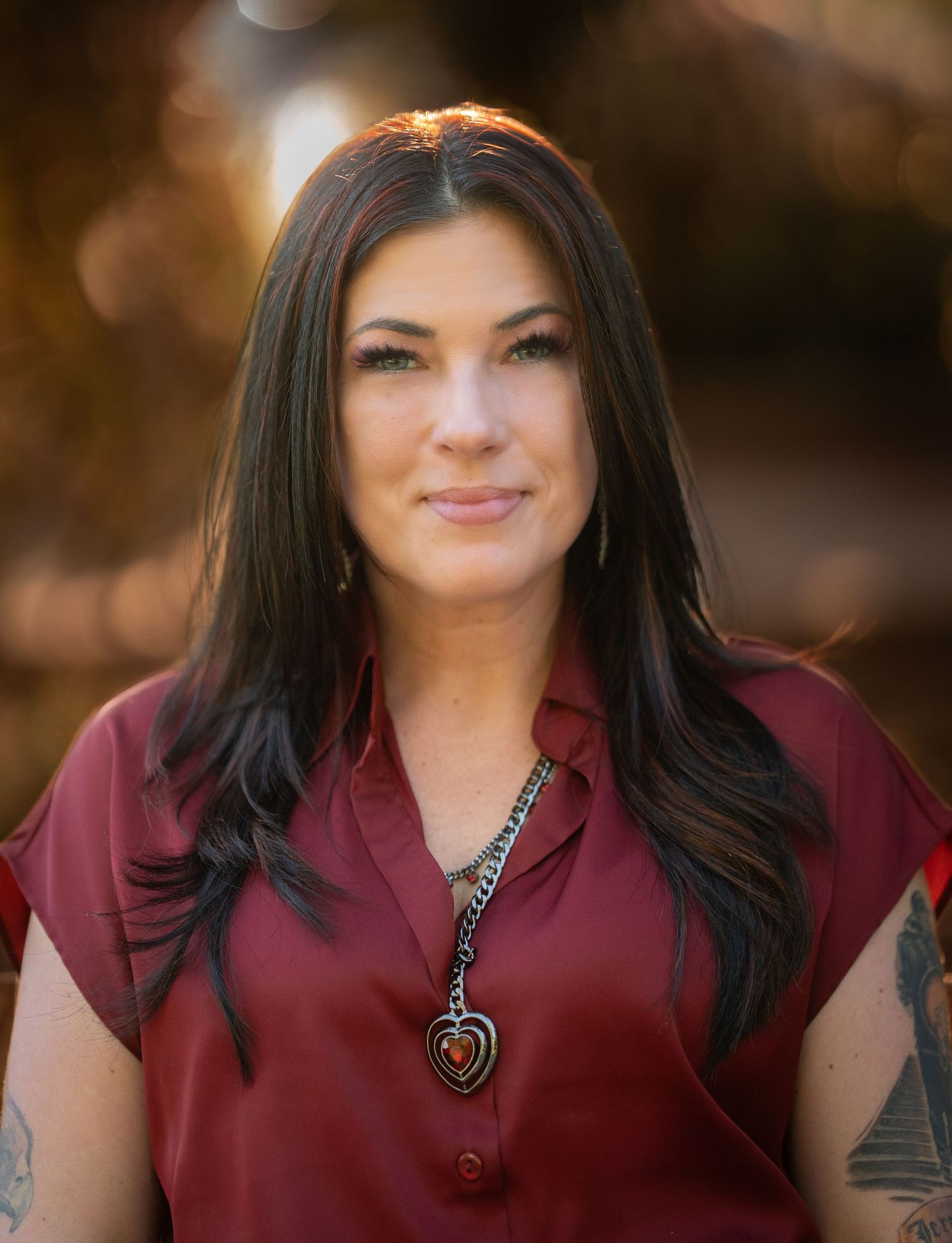 Brunette woman in a red sleeveless blouse with a heart necklace posing outdoors.