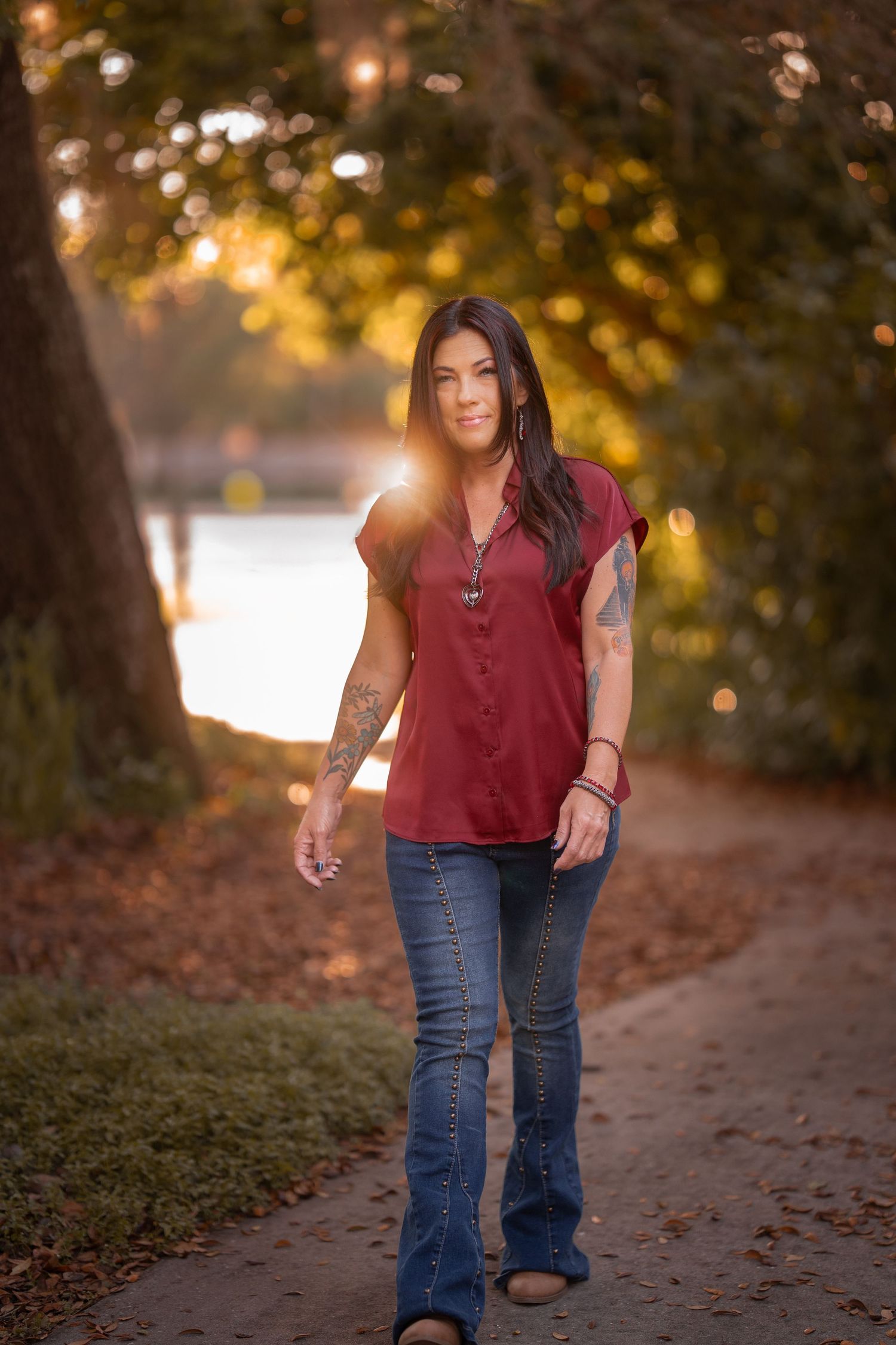 Woman with dark hair and arm tattoos in burgundy top and jeans walking on sunlit park path.