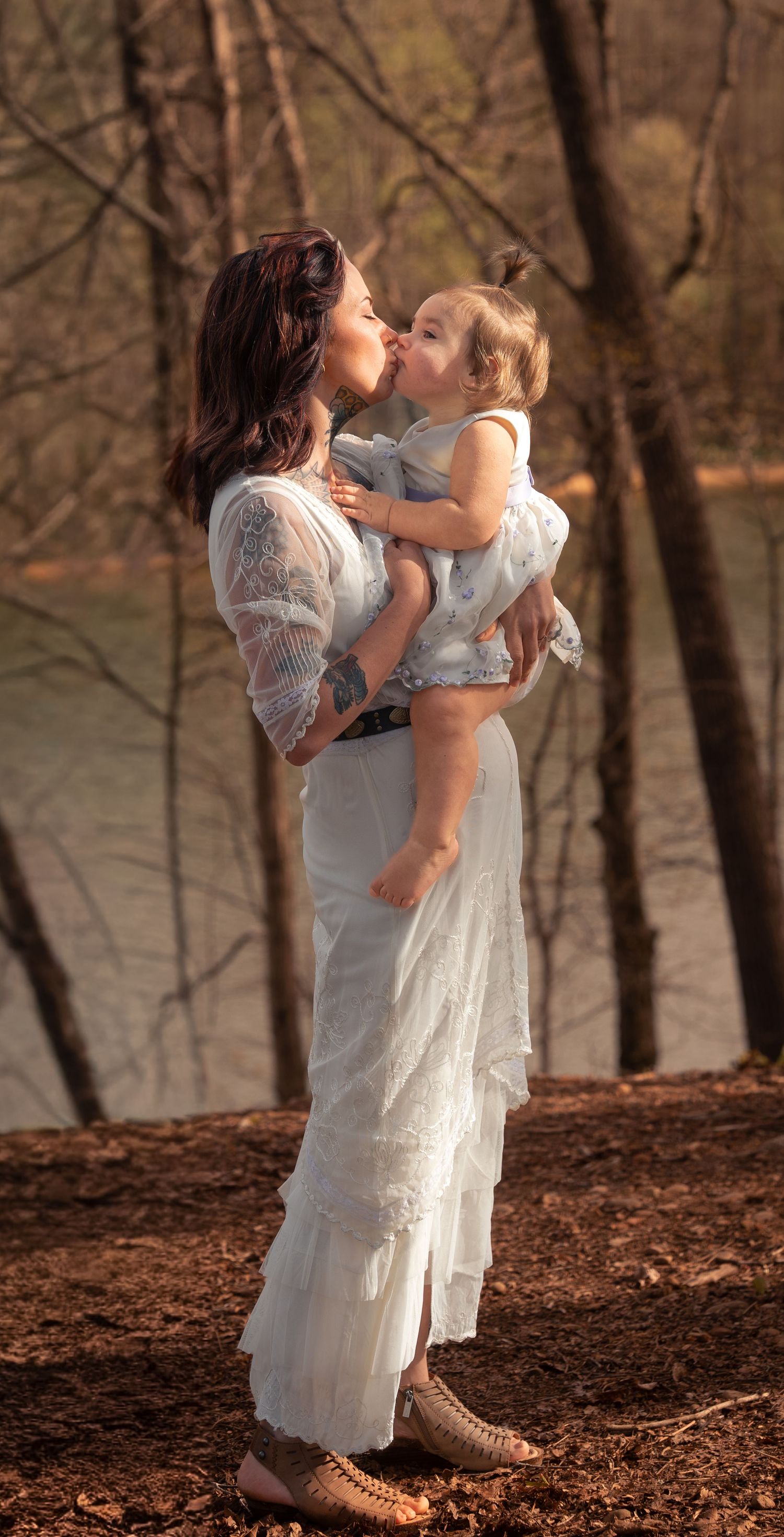 A mother in a white lace dress kisses her toddler outdoors near a lake surrounded by autumn trees.