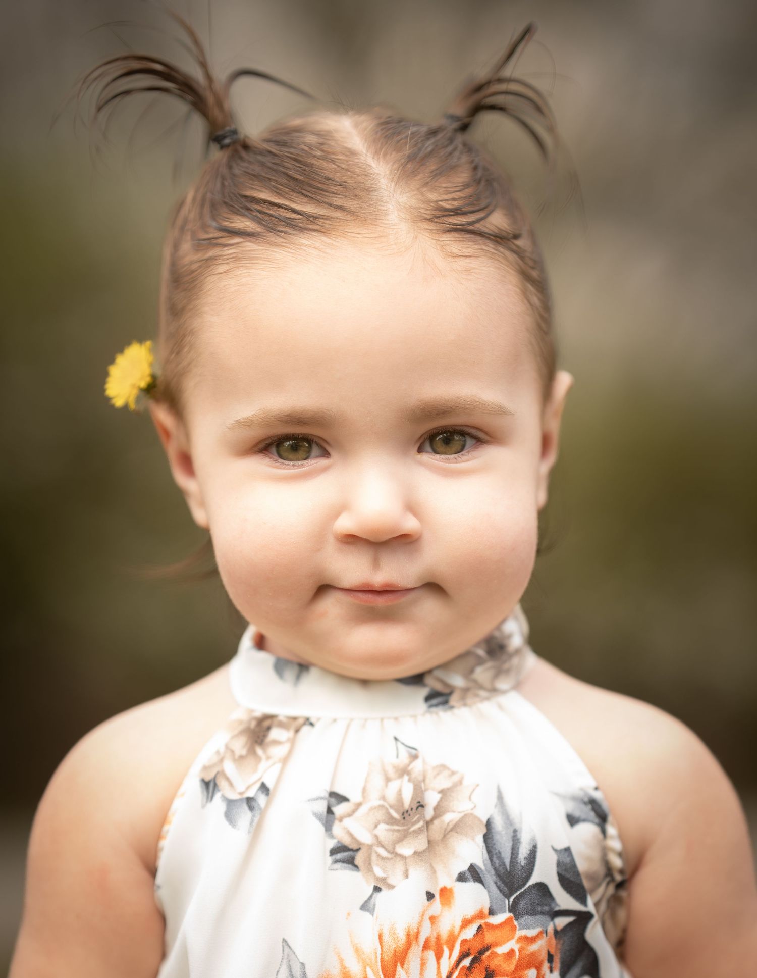 Sweet toddler girl with tiny pigtails and a yellow flower in her hair wearing a floral dress outdoors.