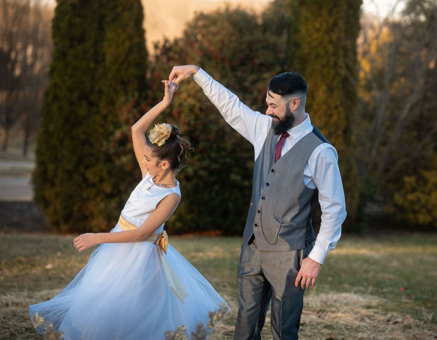 Bearded man in vest twirls young girl in blue tutu dress outdoors at sunset.