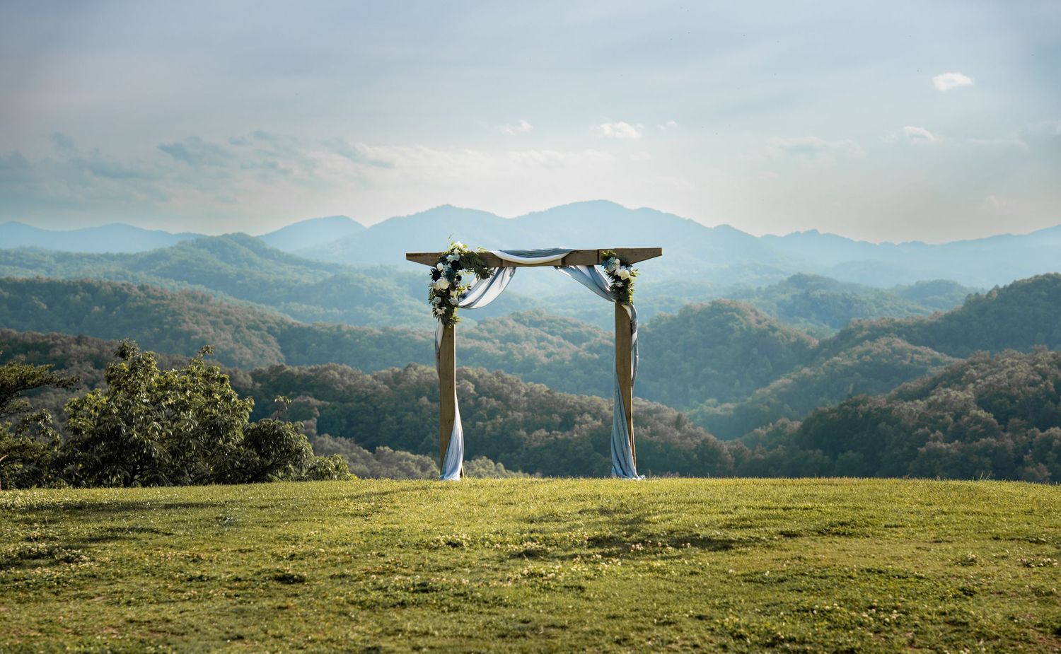 Rustic wooden wedding arch on a green hilltop with stunning mountain range backdrop under a cloudy sky.