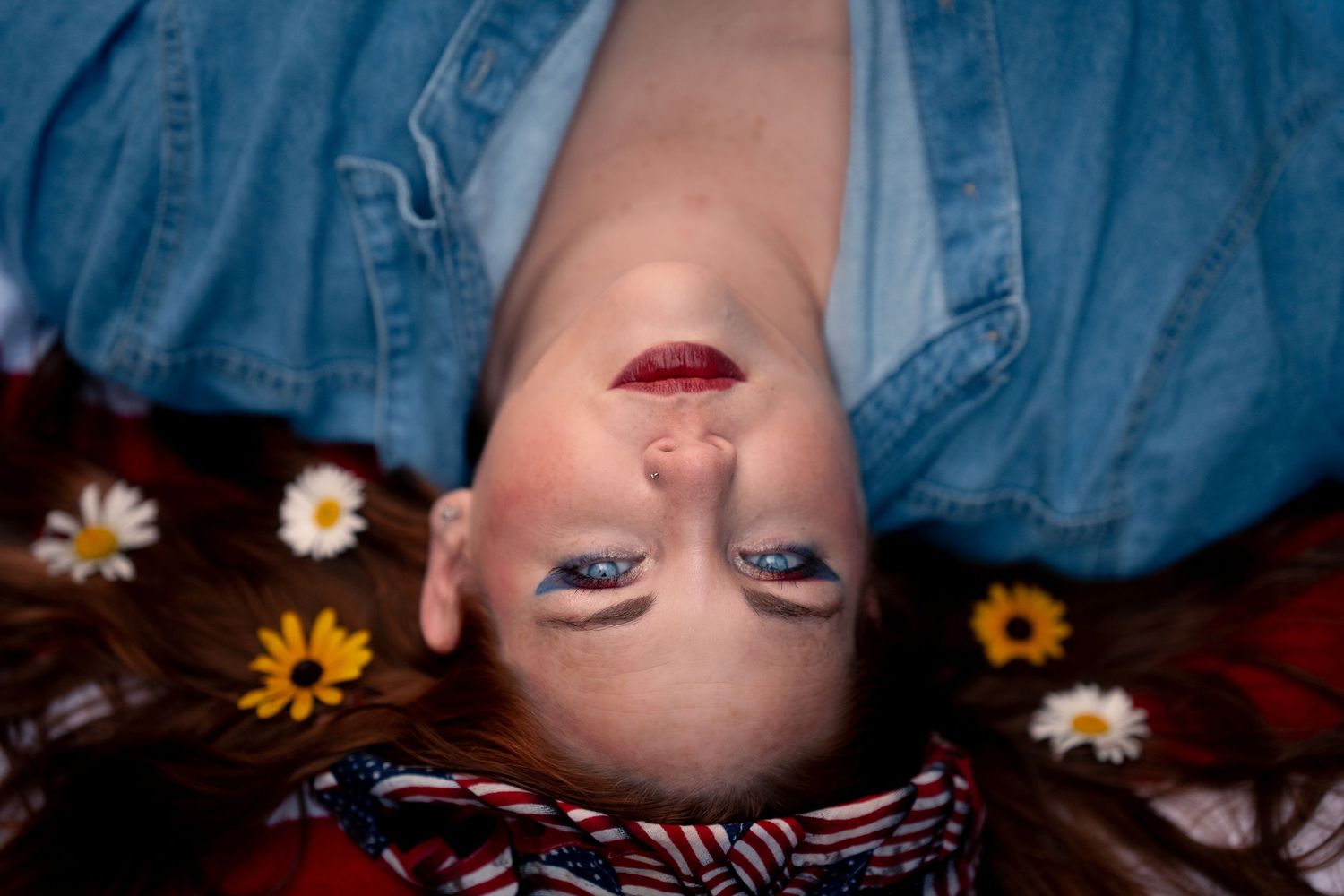 Woman lying down with flowers in hair, wearing blue eyeshadow and red lipstick, viewed from above.