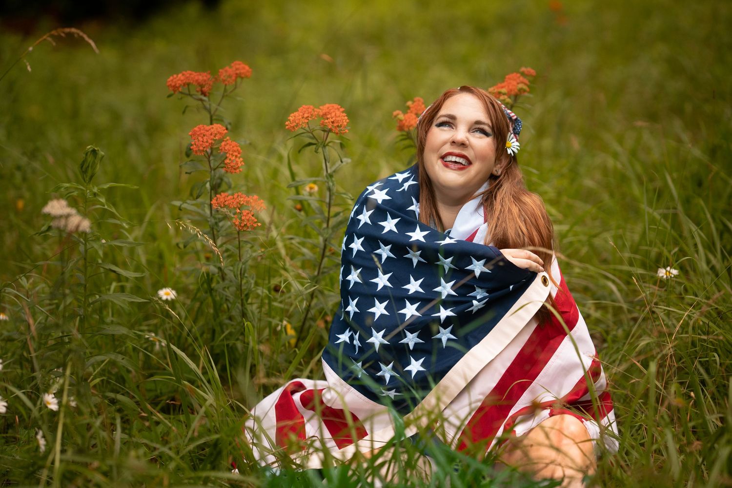 Happy redhead woman wrapped in American flag sitting in green field with wildflowers on sunny day.