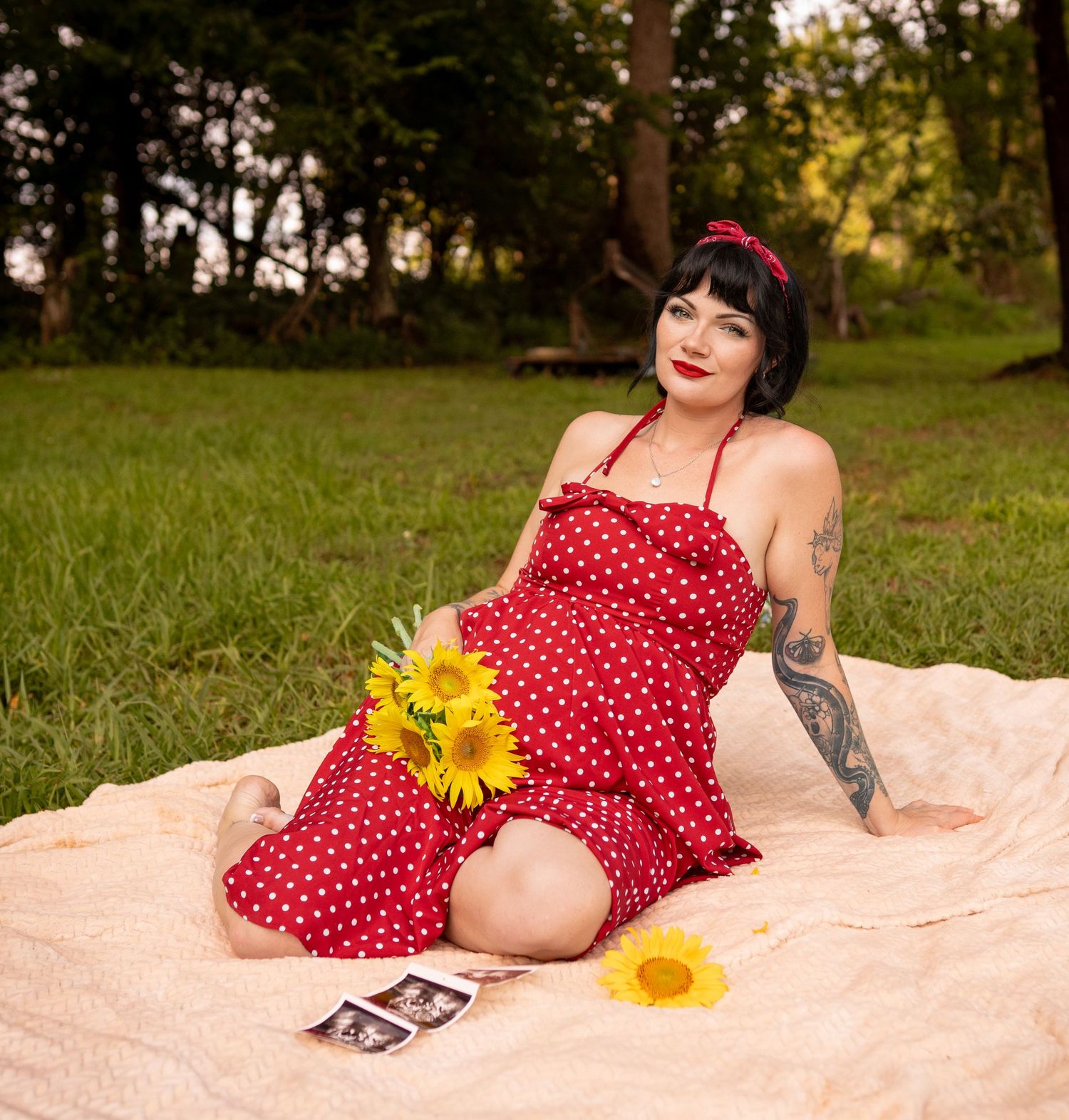 Woman in red polka dot dress holding sunflowers sits on blanket outdoors in vintage pin-up style photo.