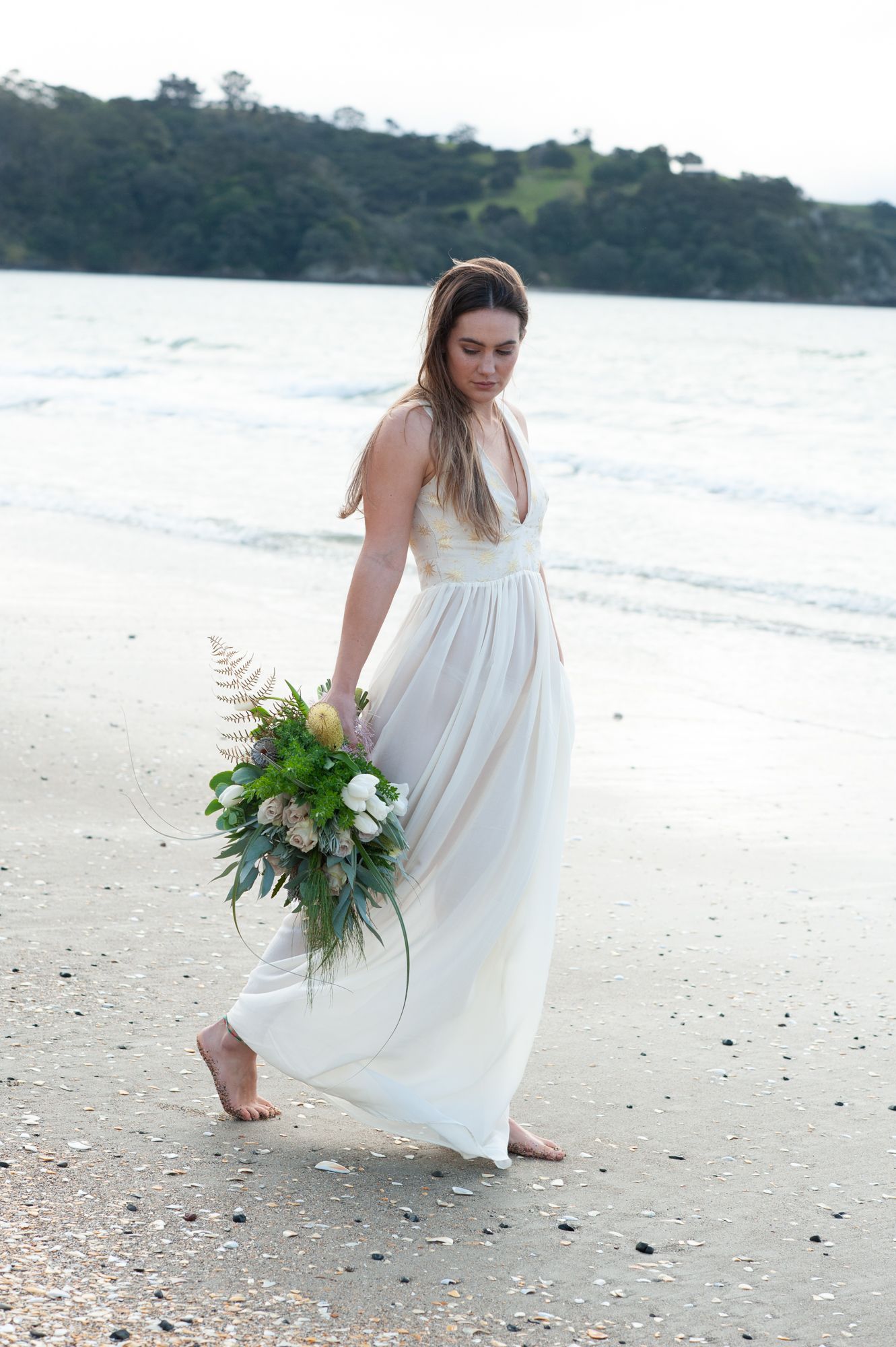 a bride walks demurely down the beach holding a beautiful bouquet of flowers hanging down by her ankles. The evening light surrounding her