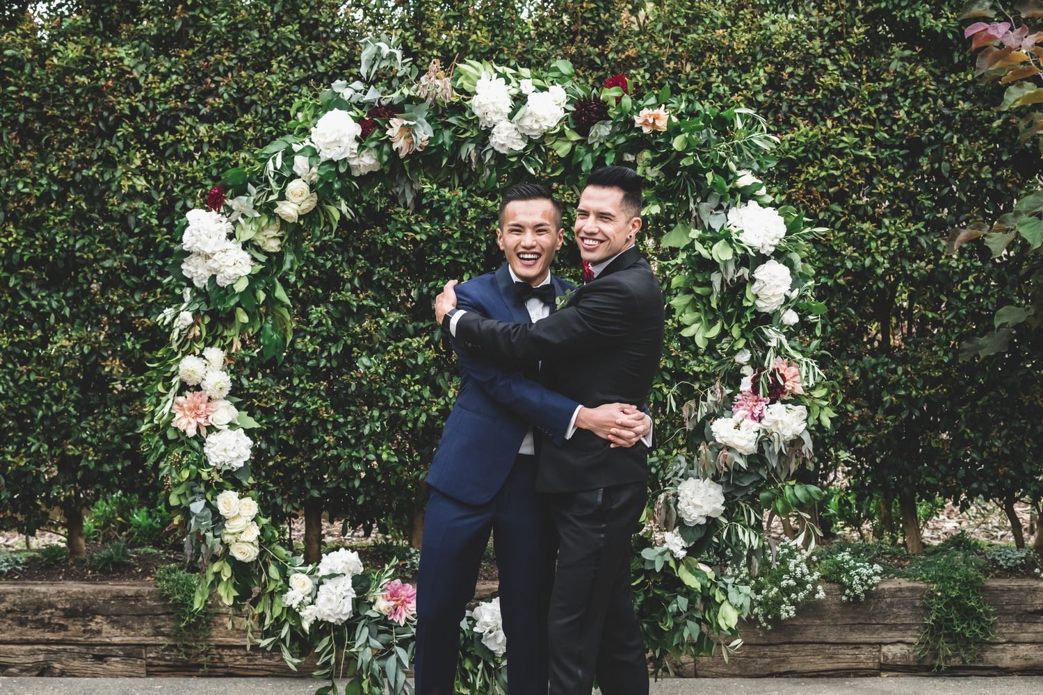 Two grooms hug each other in front of their flower circle at Mudbrick