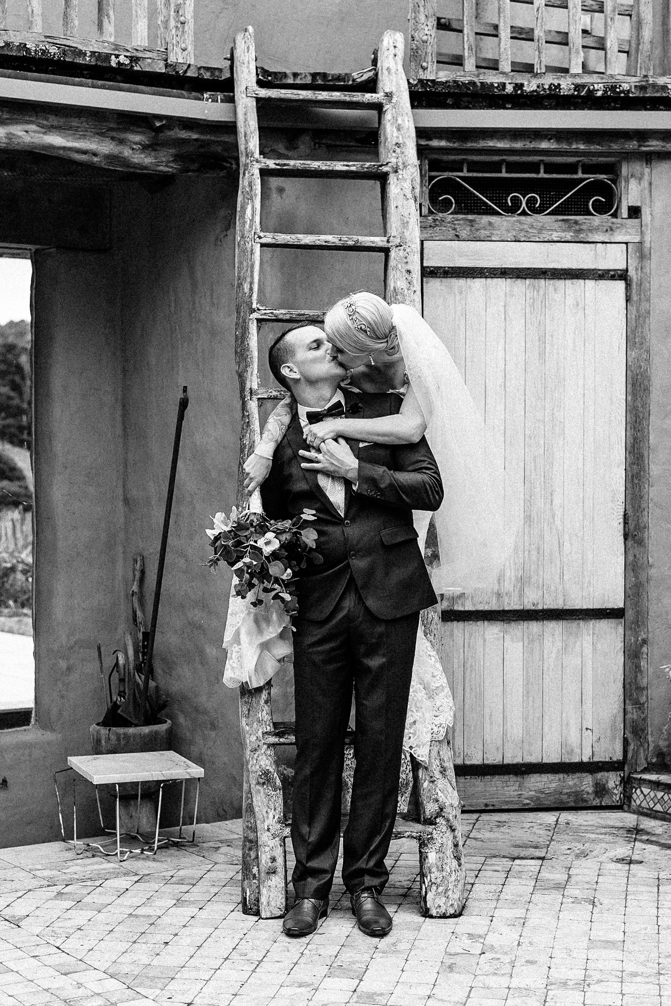 a black and white photo of a bride balanced on a ladder leaning over the groom giving him a kiss
