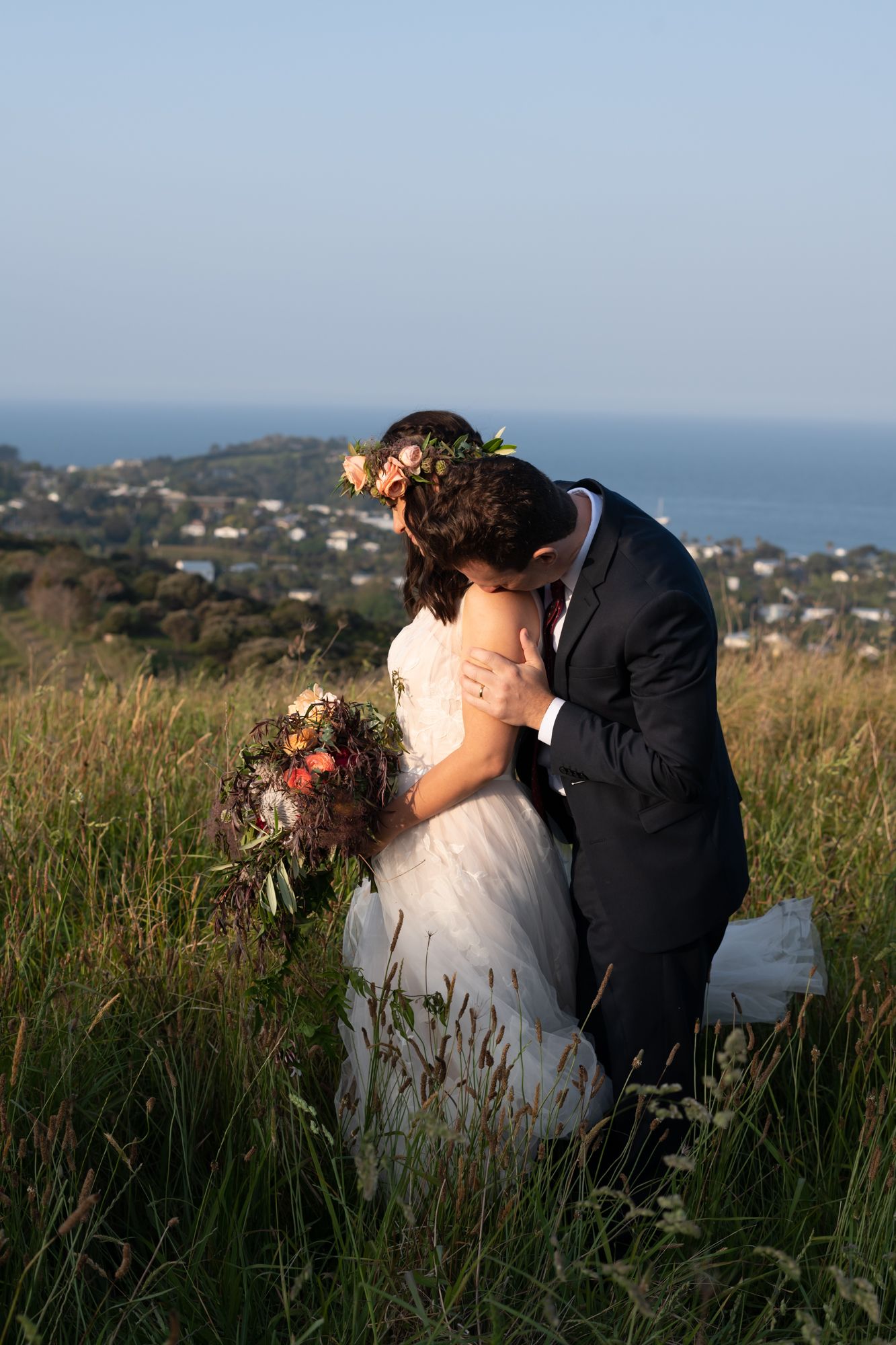 in a field of wild grass a groom affectionately kissed his brides nape of her neck in a caring affectionate embrace at the top of Mudbrick