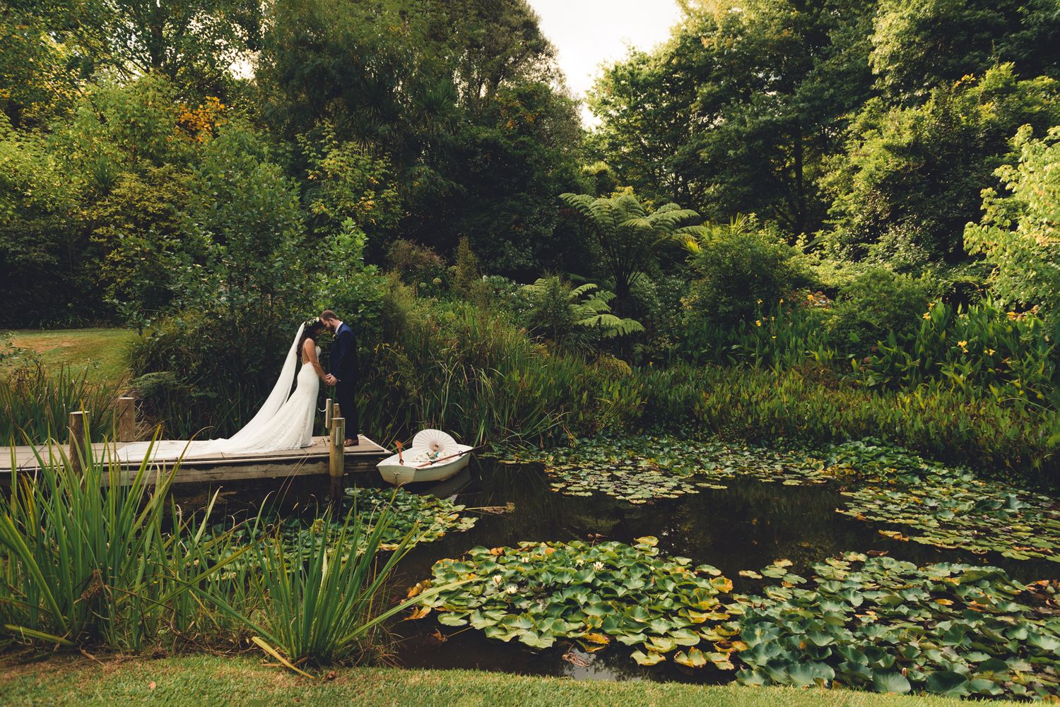 a bride and groom stand on the end of a jetty over a lake filled with Lilly pads